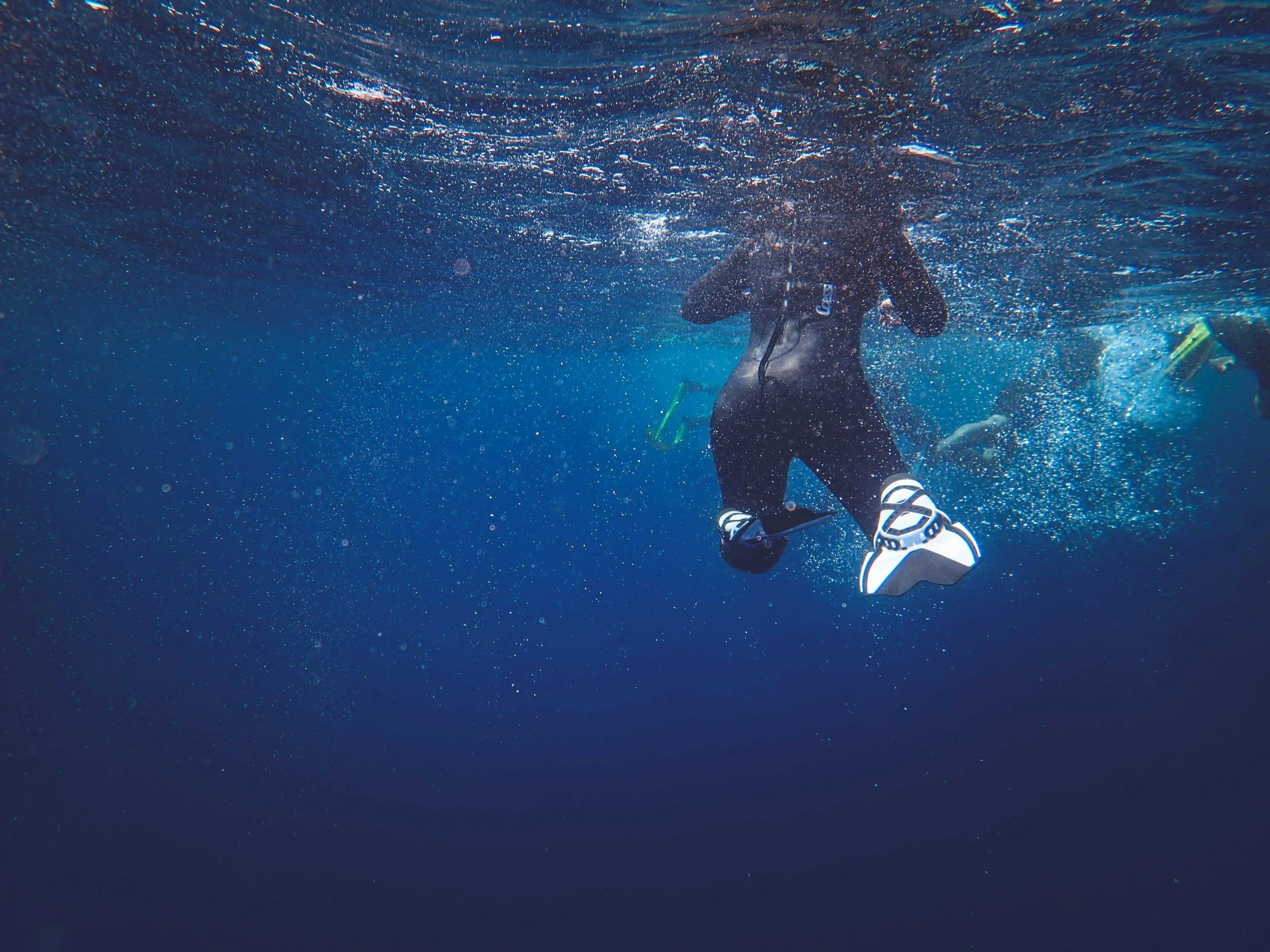 Person in wetsuit and fins swimming underwater, with bubbles surrounding them, in a deep blue ocean with other divers in the background.