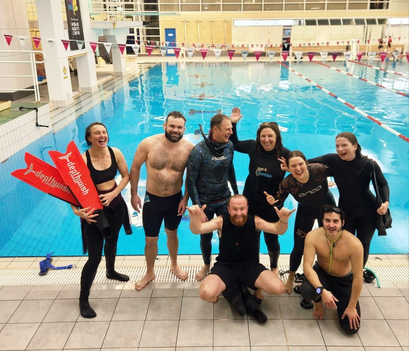 Group of people celebrating by a swimming pool, some wearing wetsuits, with one holding red fins and another sitting on the pool deck.