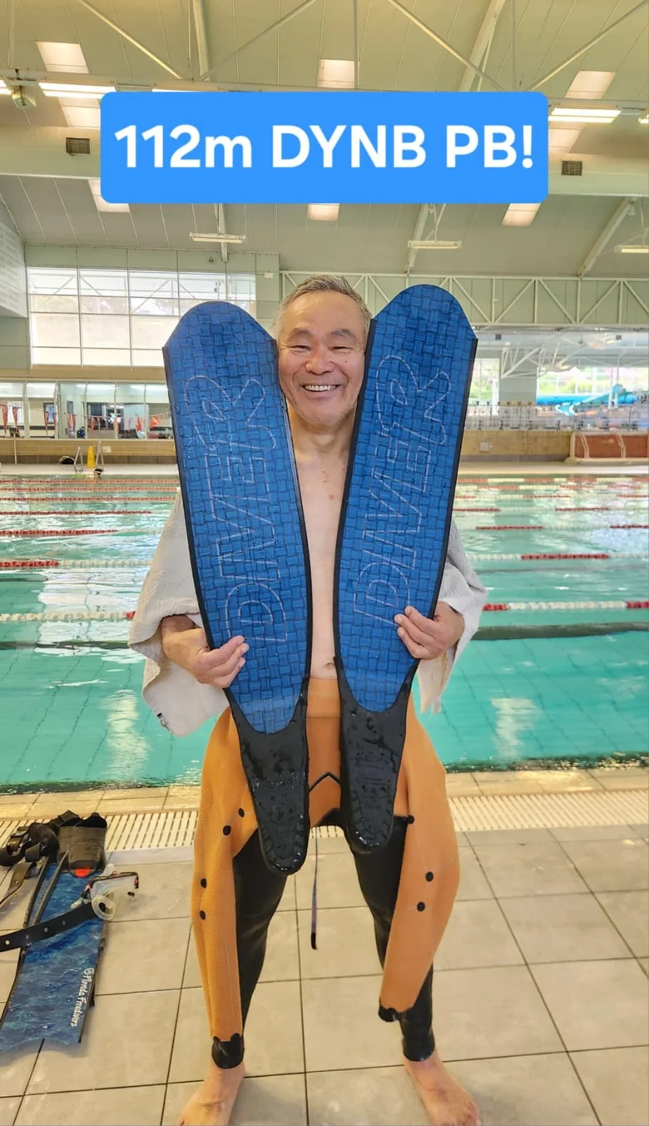 Smiling man at an indoor swimming pool holding two large blue swim fins in front of him. The pool has lane markers, and swim equipment is visible on the pool deck. The man wears tan pants and a light jacket, with a towel over his shoulders.
