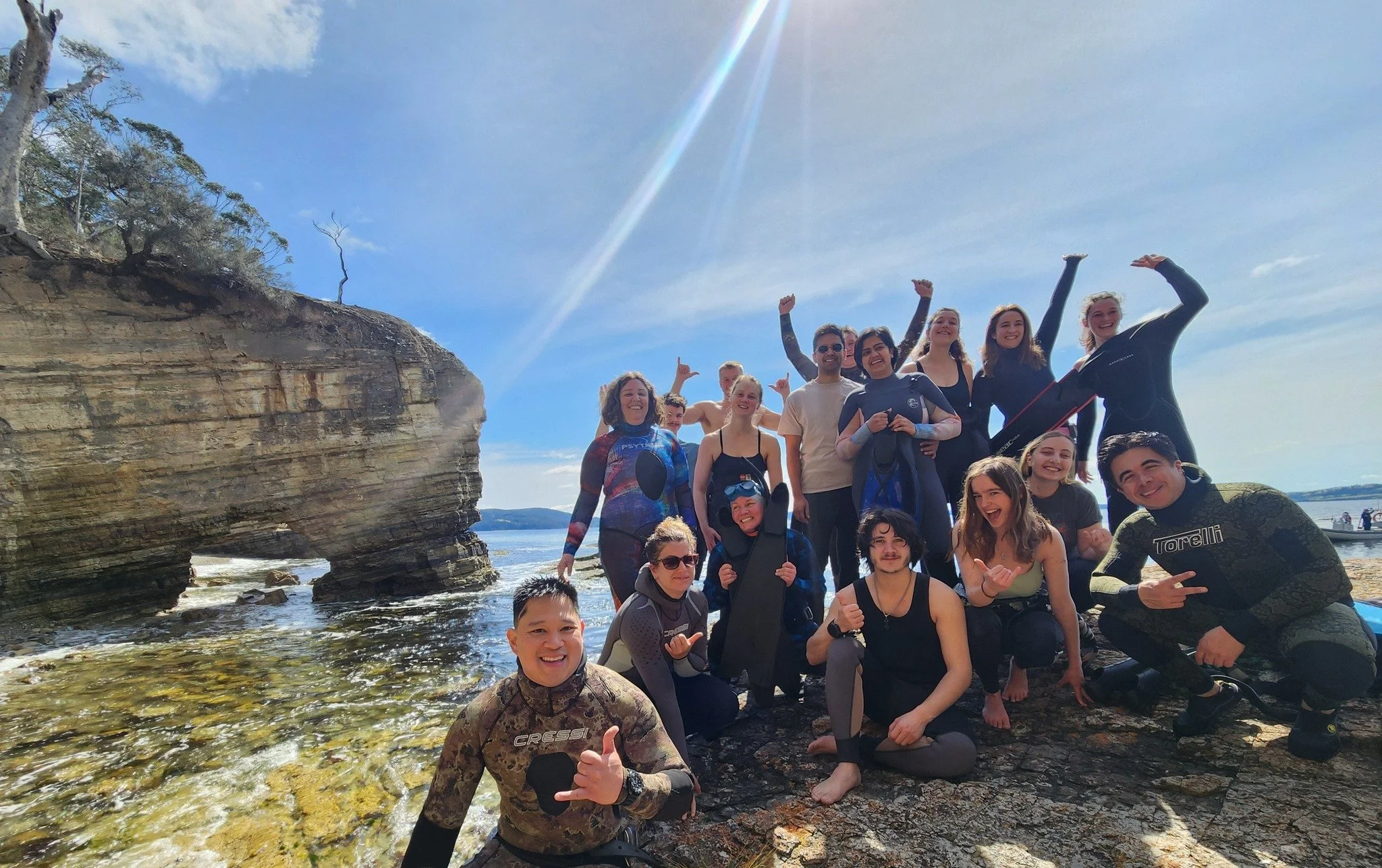 Group of people on a rocky beach, some wearing wetsuits, posing and smiling with a large rock formation and ocean in the background.