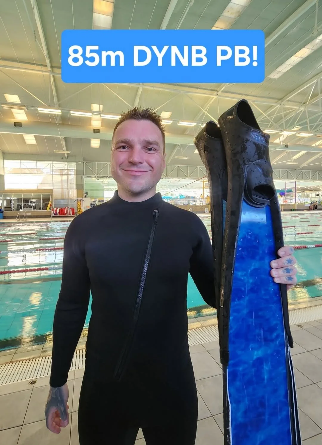 A man in a black wetsuit holding swim fins at an indoor swimming pool, with text overlay '85m DYNB PB!'.