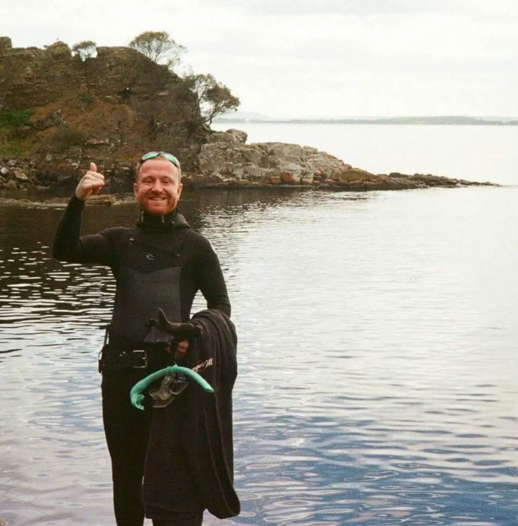 A man in scuba diving gear standing near a body of water with a rocky shoreline and hill in the background, smiling and giving a thumbs-up.