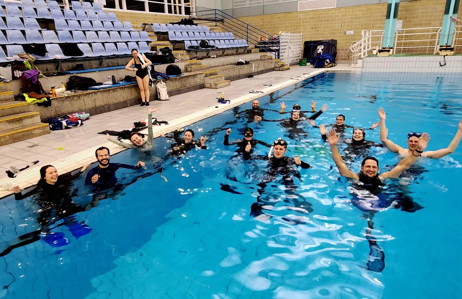 Group of people in a swimming pool celebrating, with some raising their hands and others smiling, as a woman on the poolside prepares gear.