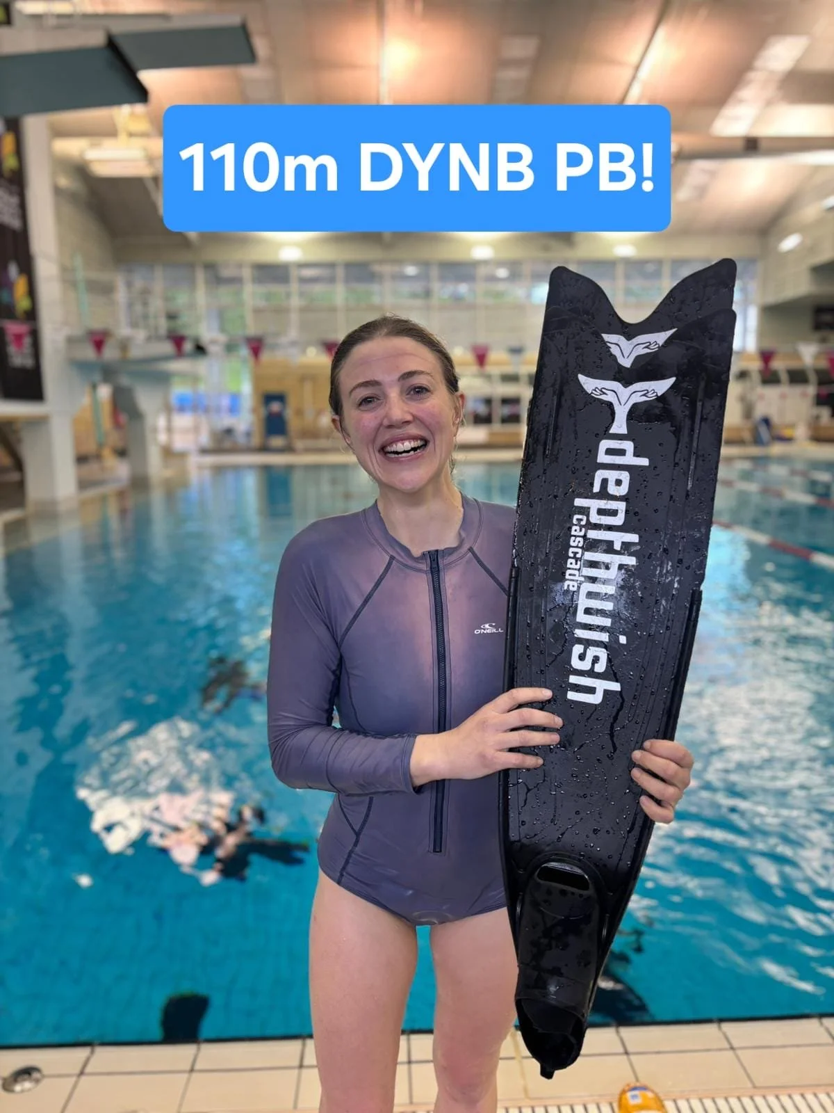 A woman standing next to a swimming pool, holding a black kickboard with the words 'Apephus, Pasadena' on it, and smiling at the camera. She is wearing a gray swim jacket, and there are swimmers in the pool behind her.
