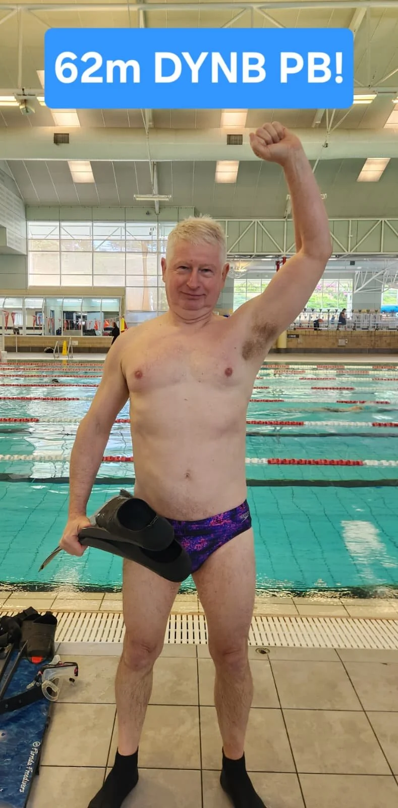 An elderly man standing at an indoor swimming pool, holding swim fins in one hand, and raising his arm in a fist pump gesture with a blue sign overhead that reads '62m DYNB PB!'
