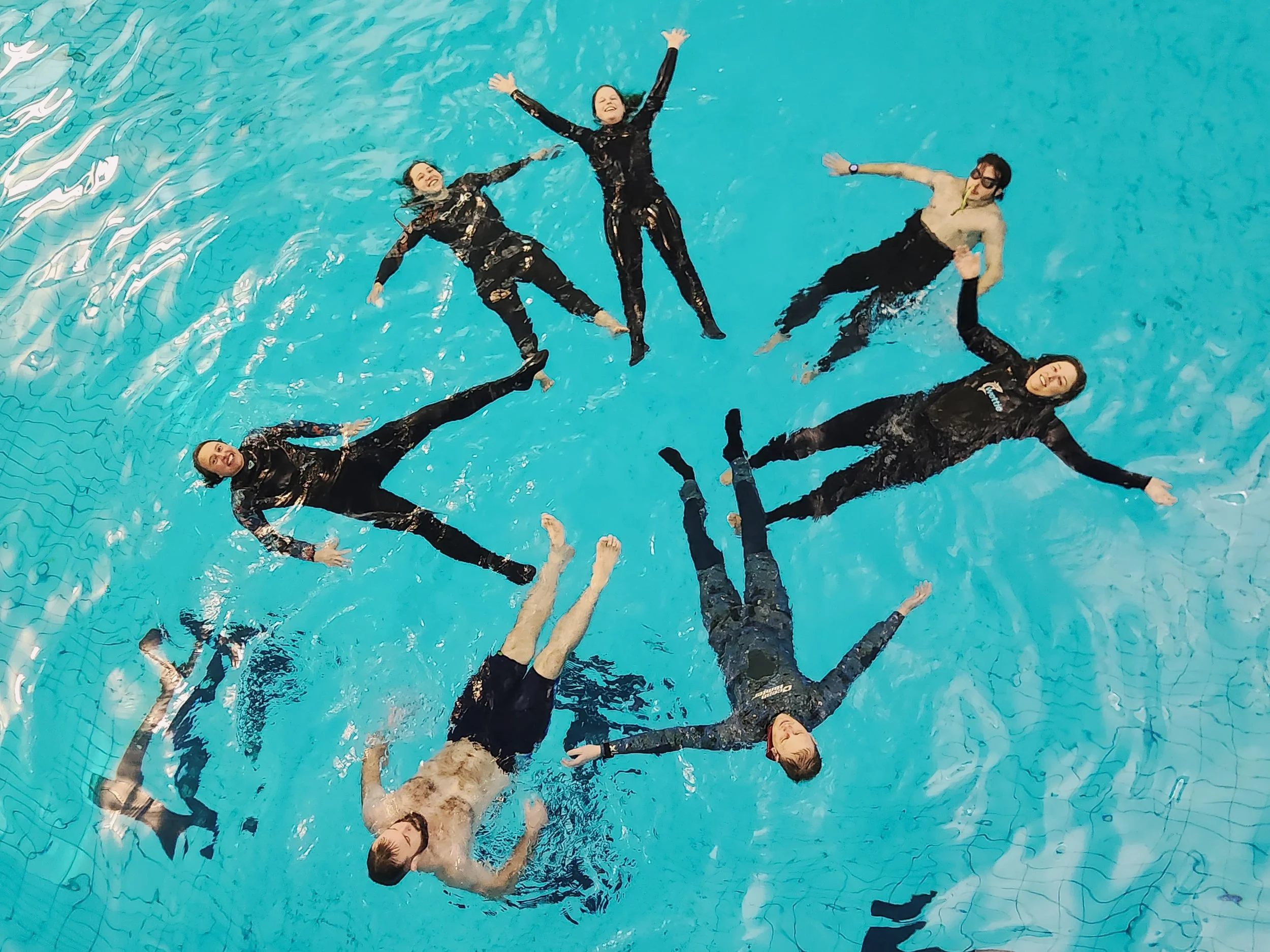 Group of eight people floating in a swimming pool with arms and legs spread out creatively, wearing swimwear and casual clothing, with one person in glasses.