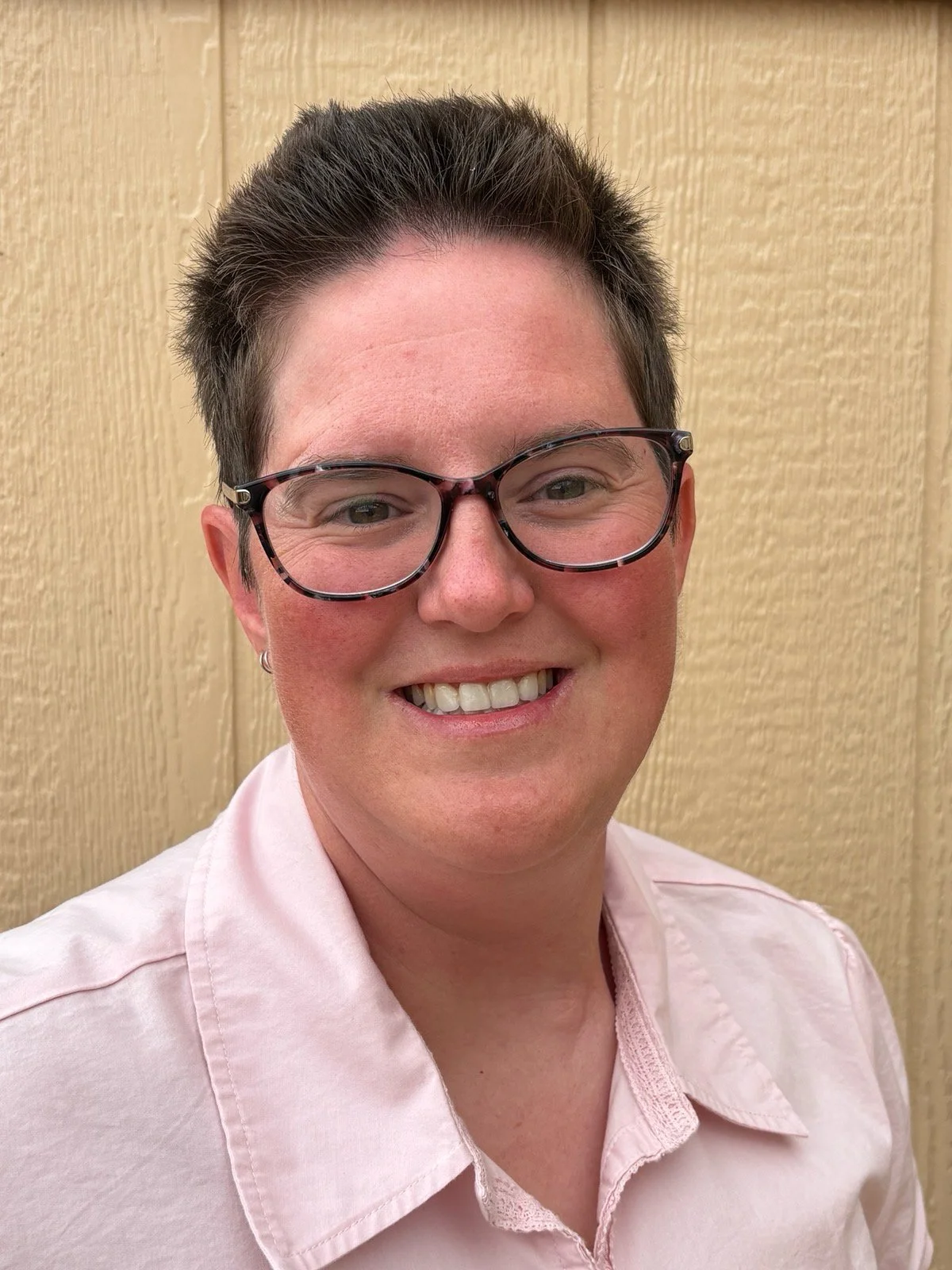 A person with short dark hair, wearing glasses and a light pink collared shirt, smiling in front of a wooden wall background.