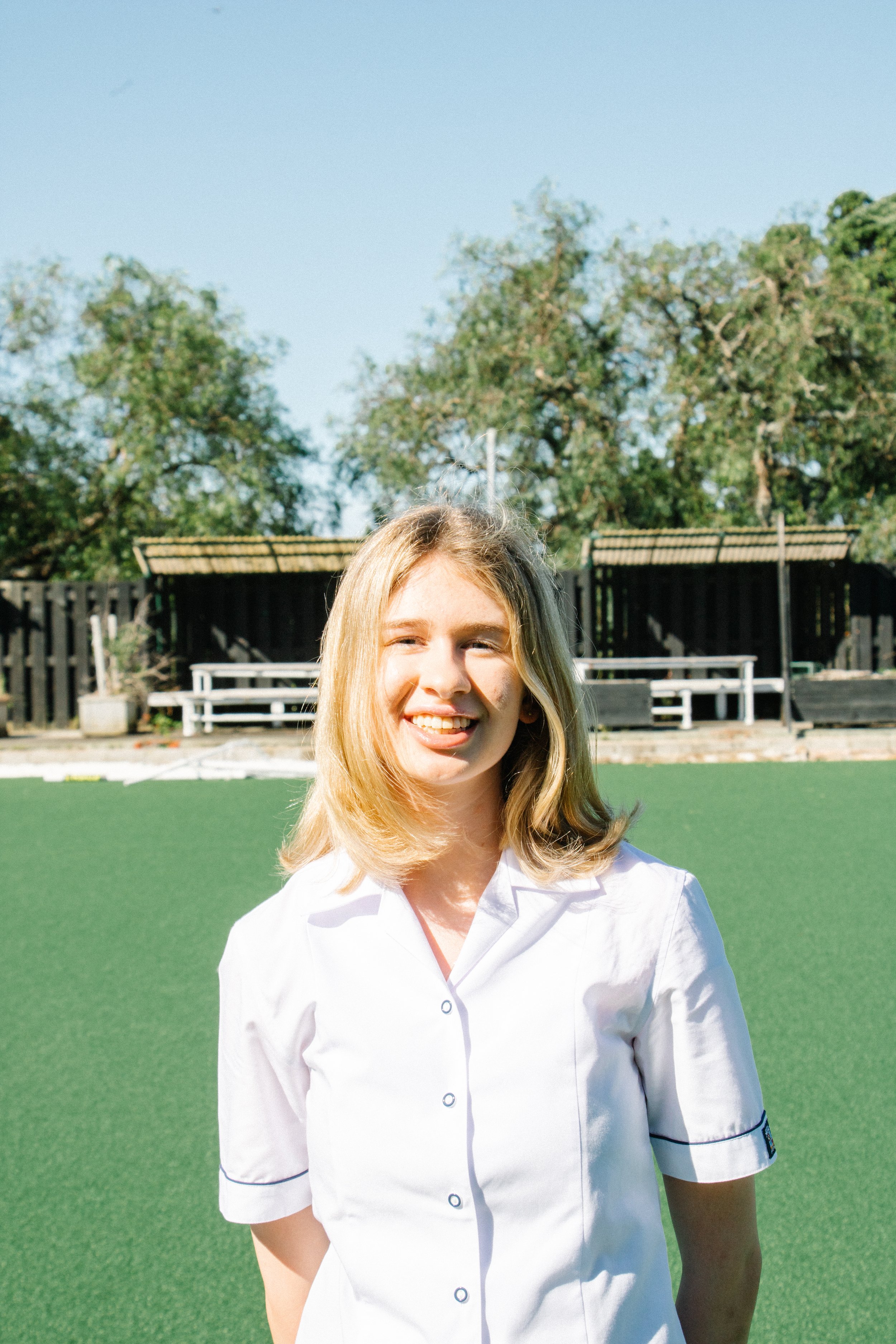 Young woman with blonde hair smiling outdoors on a sunny day, wearing a white shirt, standing in front of a green field with trees and wooden structures in the background.