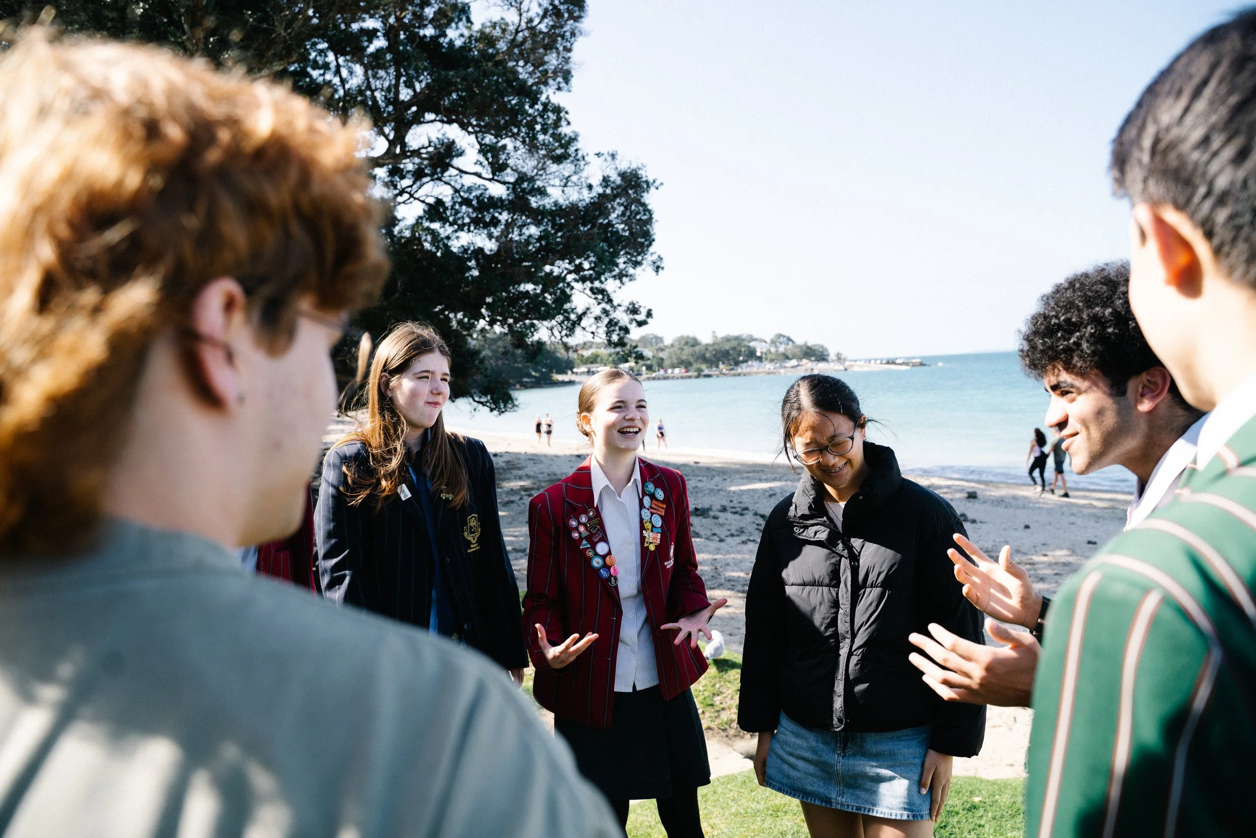 Group of diverse teenagers standing on a grassy area near a beach, socializing and laughing on a sunny day.
