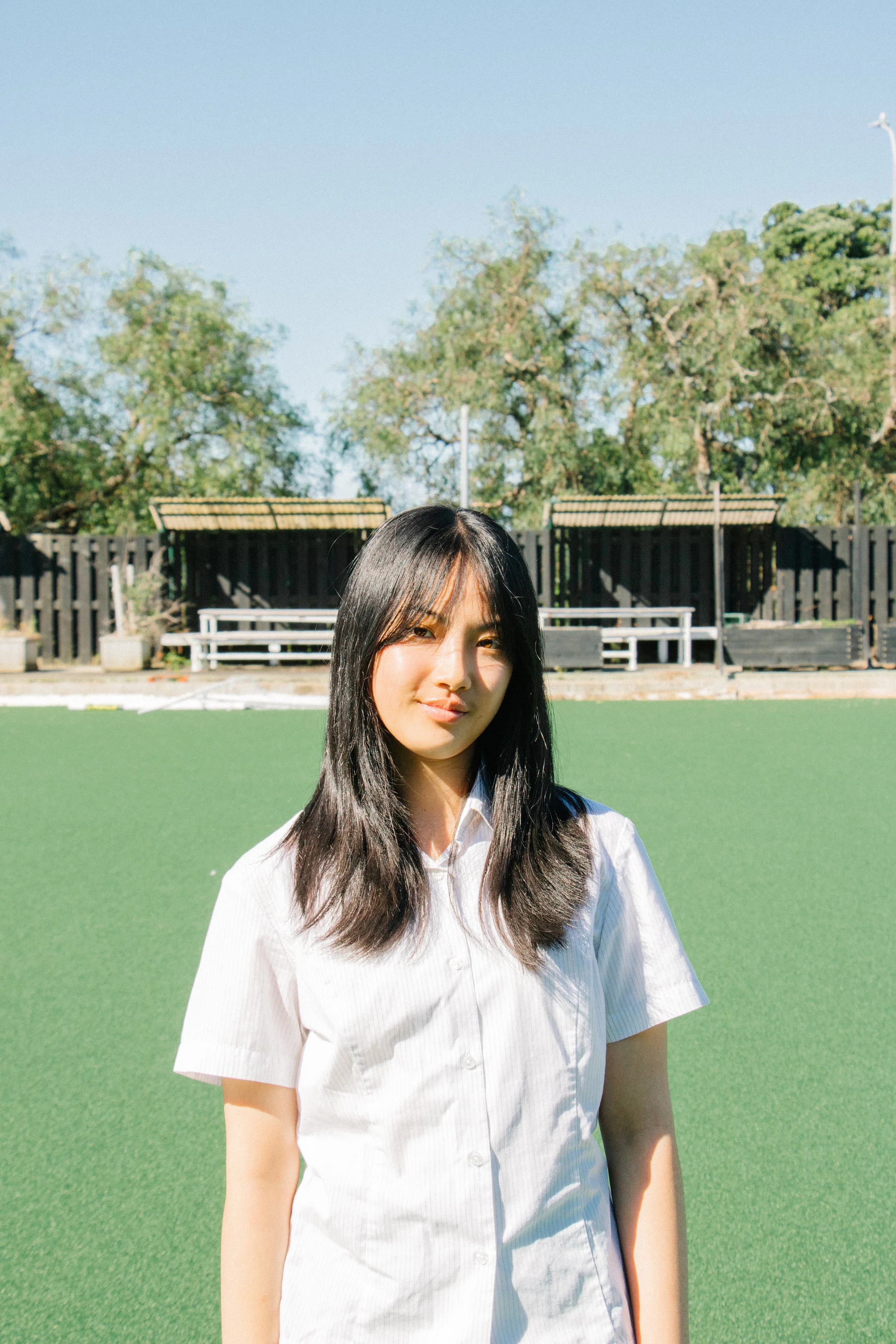 Young woman with long black hair wearing a white shirt standing outside on a sunny day. Green grass field and trees in the background.