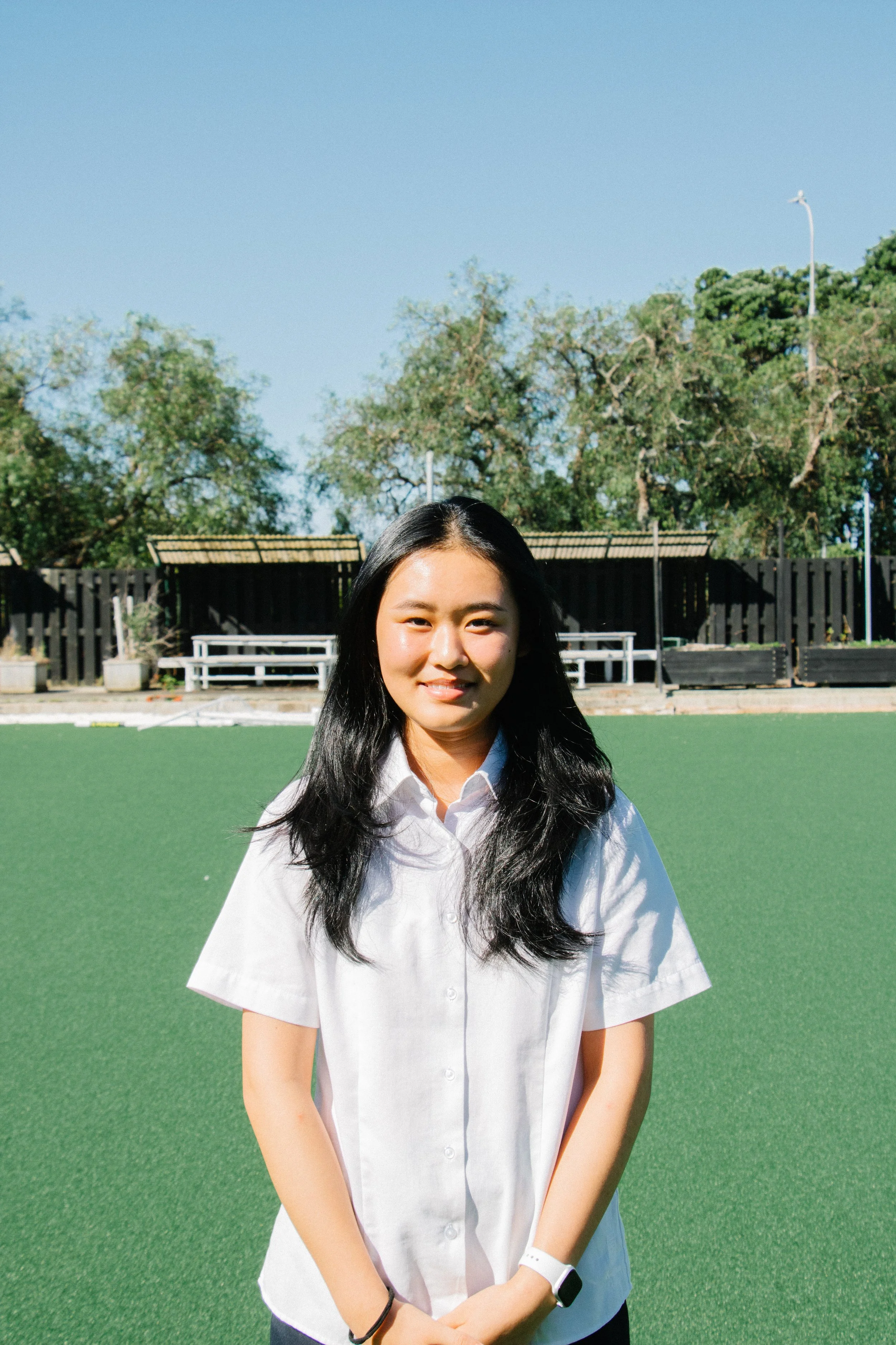 Young woman with long black hair, wearing a white short-sleeve shirt, standing on a green sports field on a sunny day.