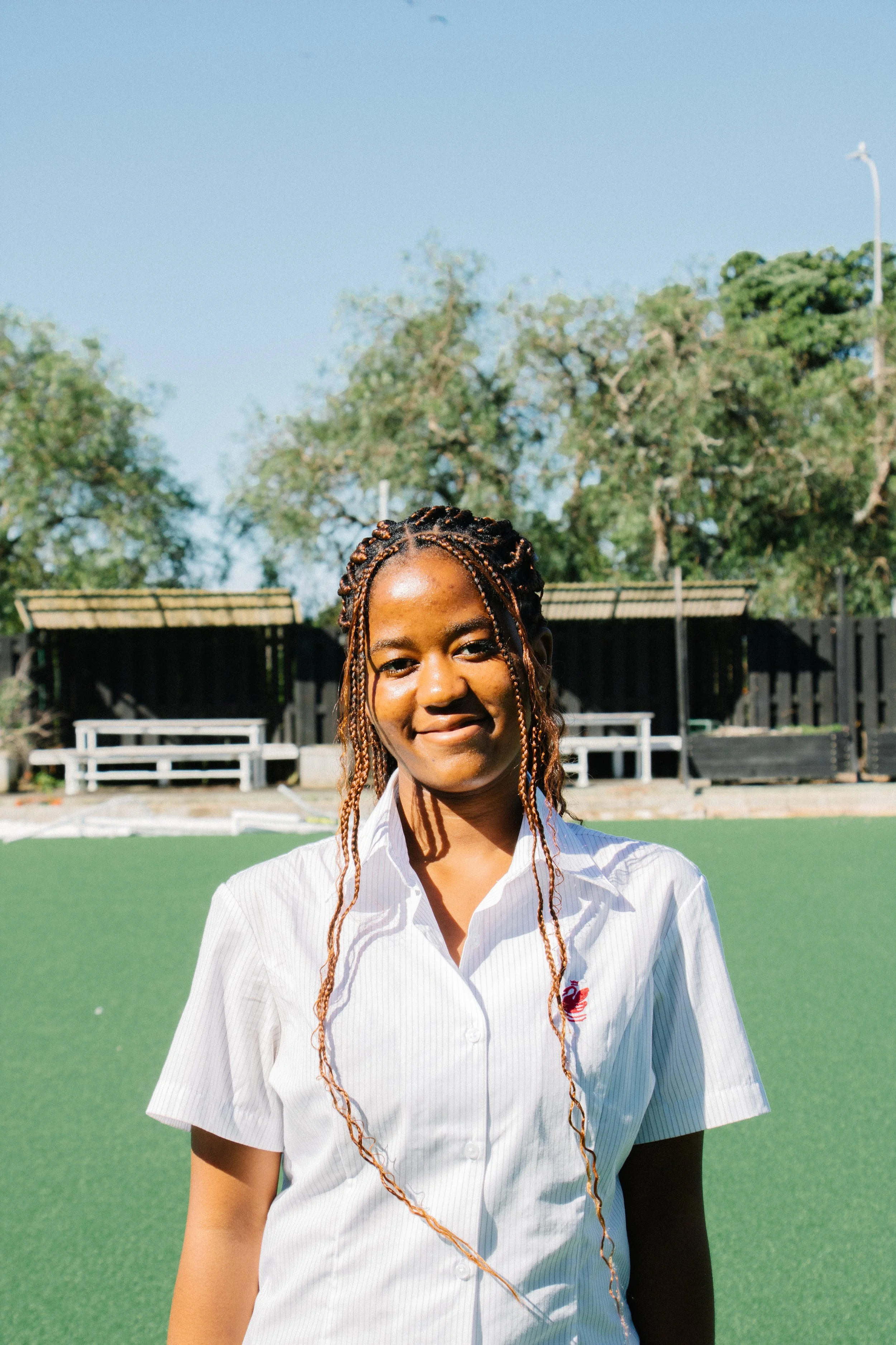 A young woman with braided hair is standing outdoors on a grassy field, smiling at the camera, with trees and benches in the background.