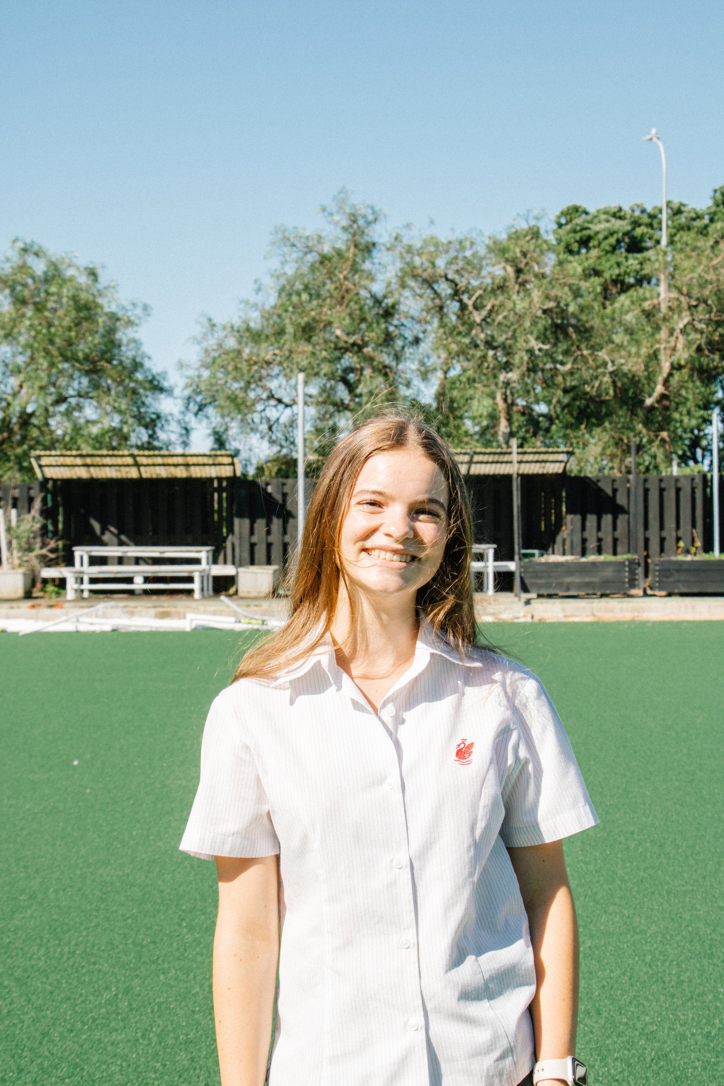 A young woman with long blonde hair smiling outdoors on a sunny day, wearing a white short-sleeved blouse, standing on a green artificial turf field with trees and black fences in the background.