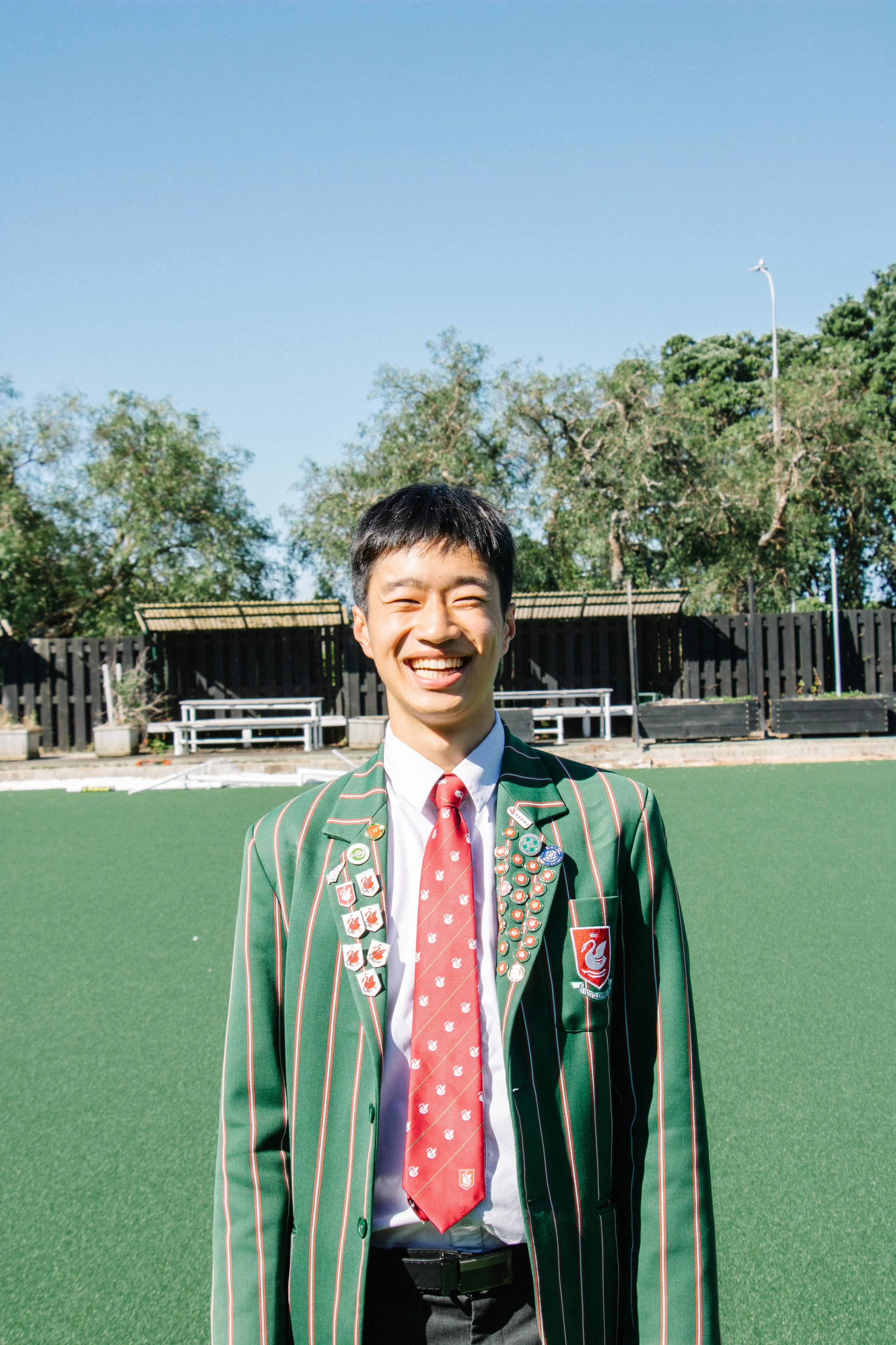 A young man standing outdoors on a grassy field, wearing a green blazer with white and red vertical stripes, a white shirt, a red tie, and an array of pins and badges on their blazer. The background includes trees, a black fence, and a clear blue
