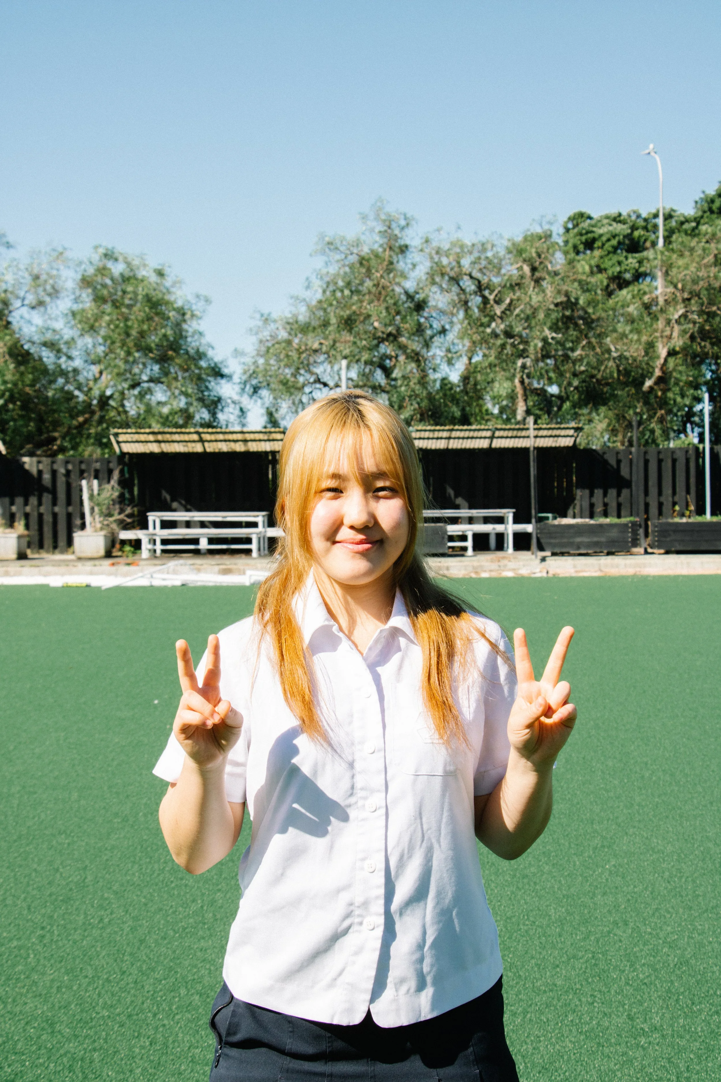 A young woman with blonde hair smiling and making peace signs with both hands on a green outdoor field.