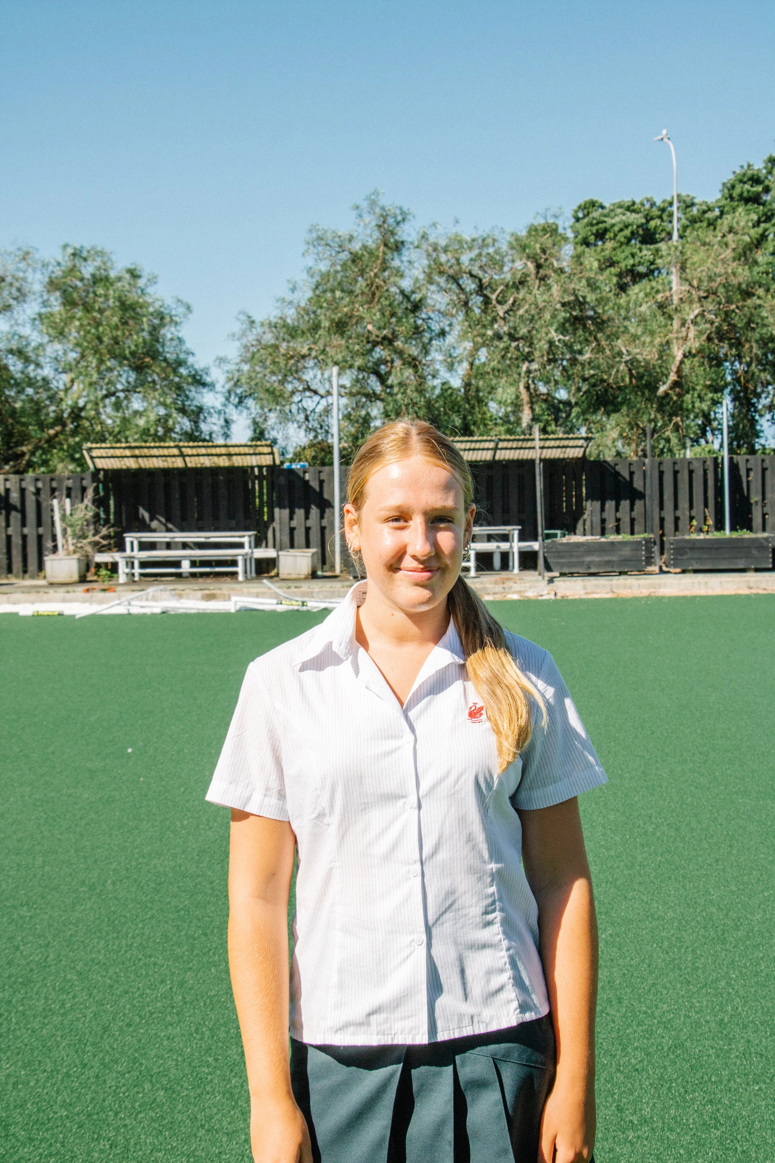A young girl with long blonde hair tied in a ponytail, wearing a white short-sleeved shirt and gray shorts, standing on a green sports field with trees and benches in the background.