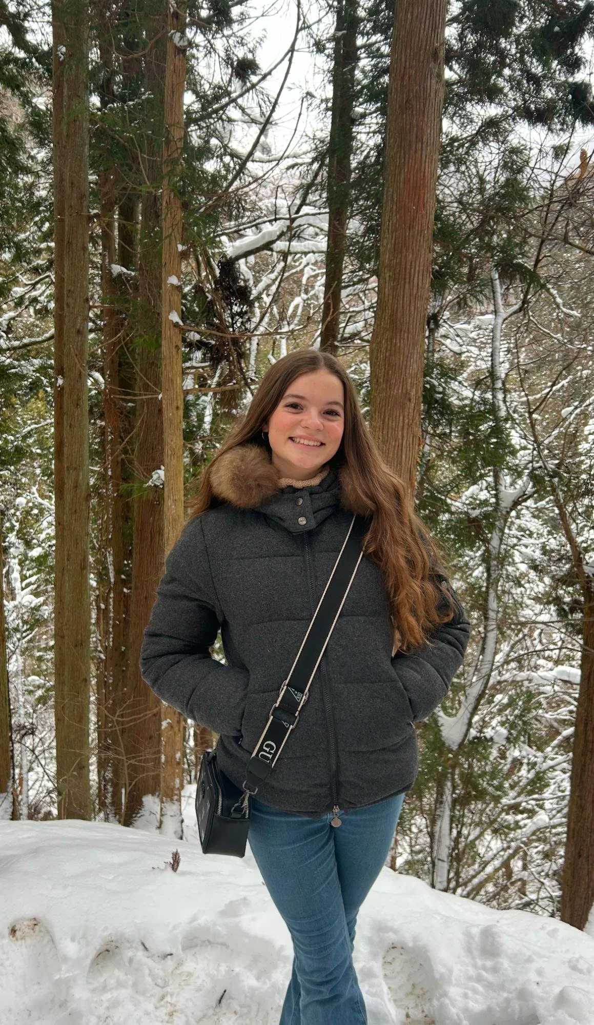 A young woman with long brown hair smiling outdoors in a snowy forest, wearing a black winter jacket with fur collar and blue jeans.