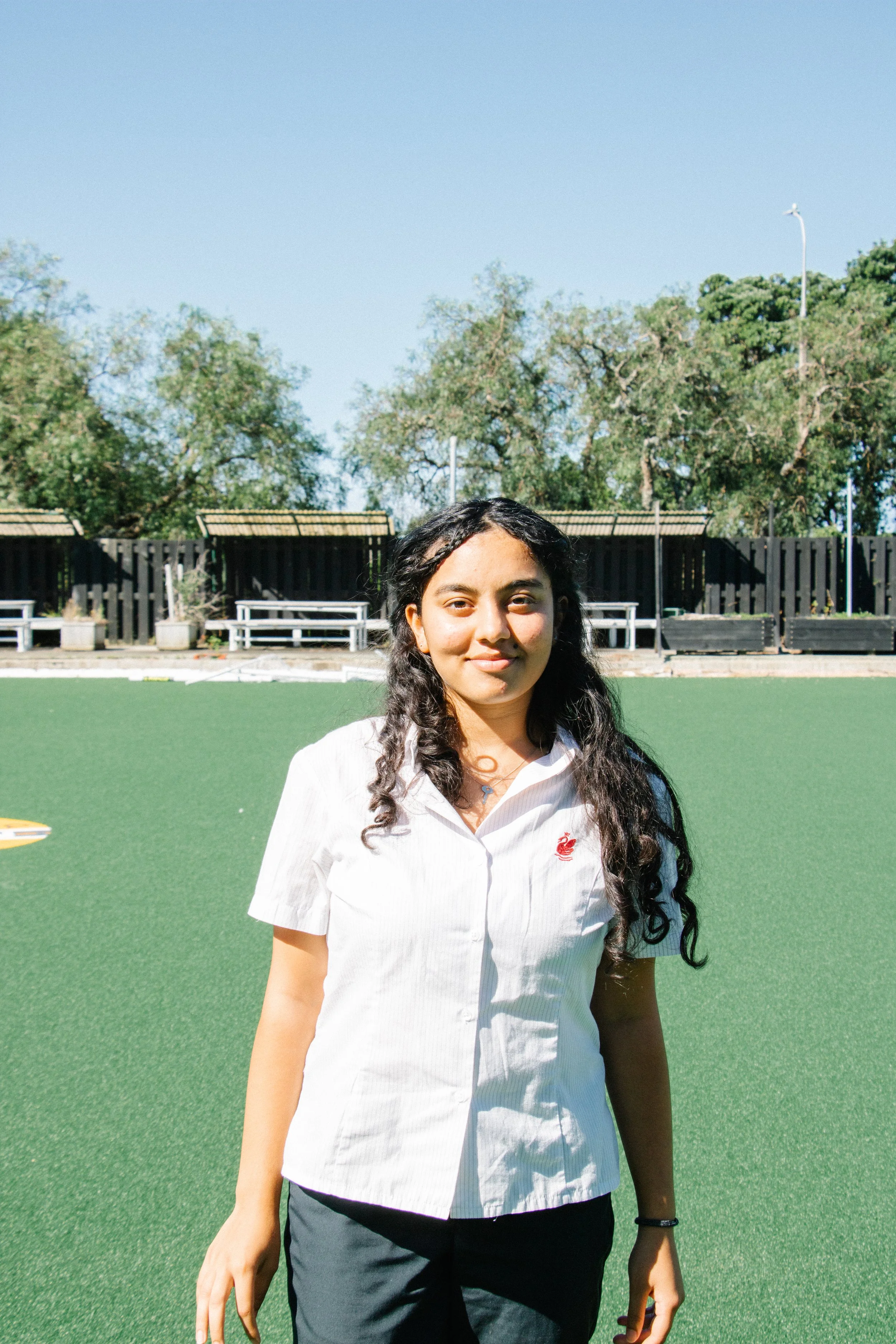 A young woman with long, curly black hair standing outdoors on a green field with trees, benches, and a black fence in the background, wearing a white short-sleeved shirt and black pants.
