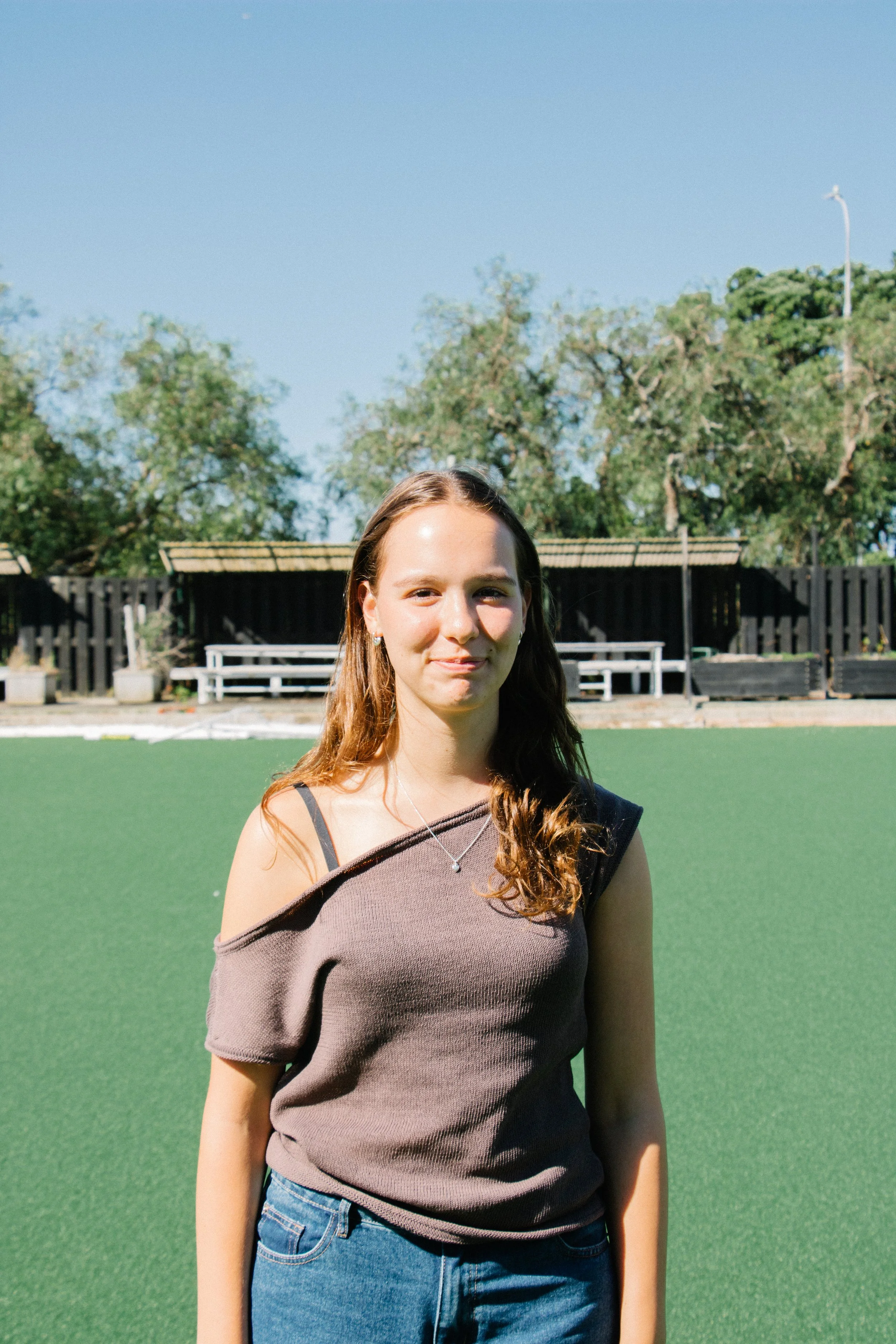 Young woman standing on a green field outdoors, wearing a brown top and jeans, with trees and a fence in the background on a sunny day.