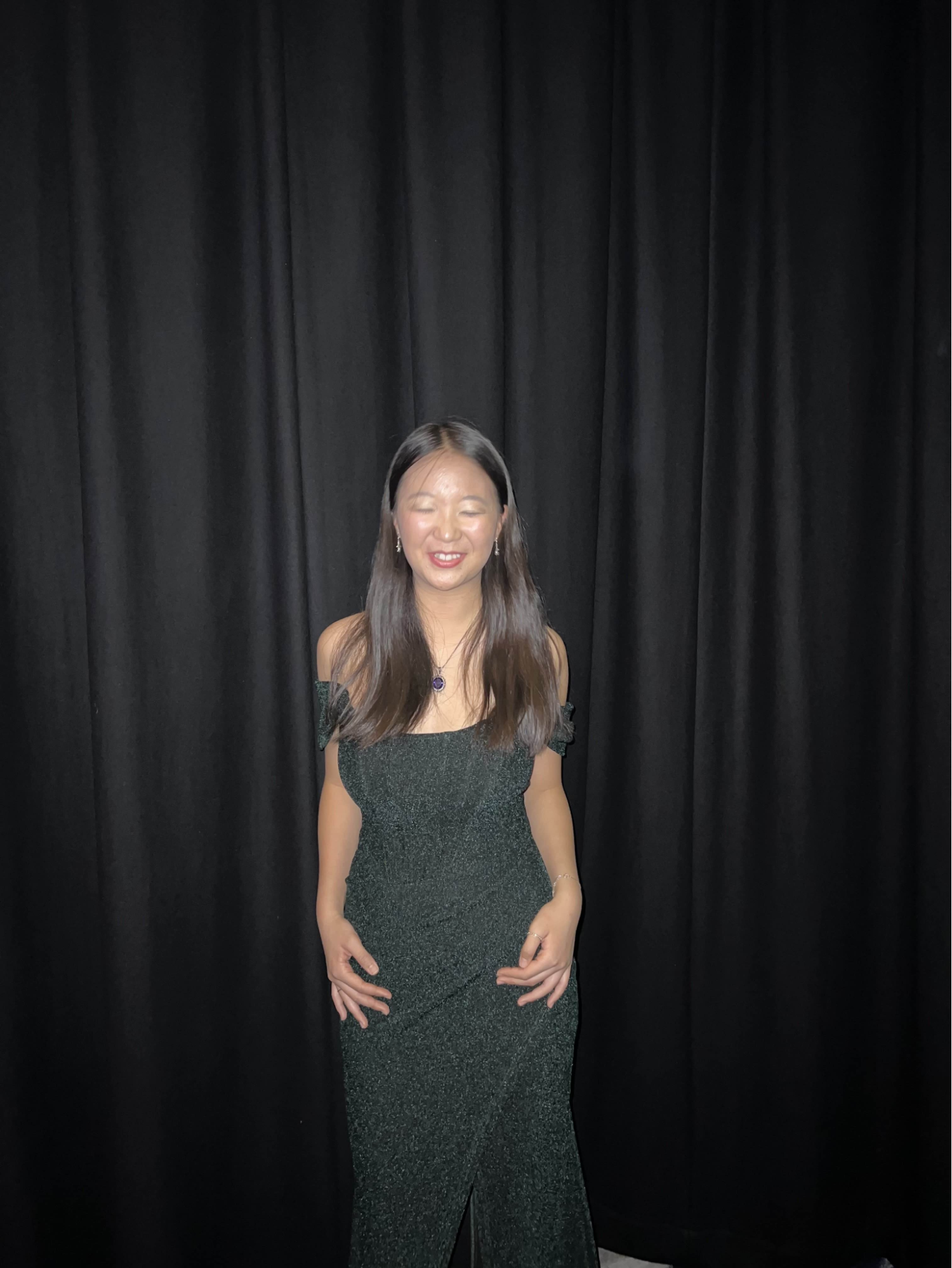 A woman with long dark hair wearing an off-shoulder black dress standing in front of black curtains, smiling with eyes closed.