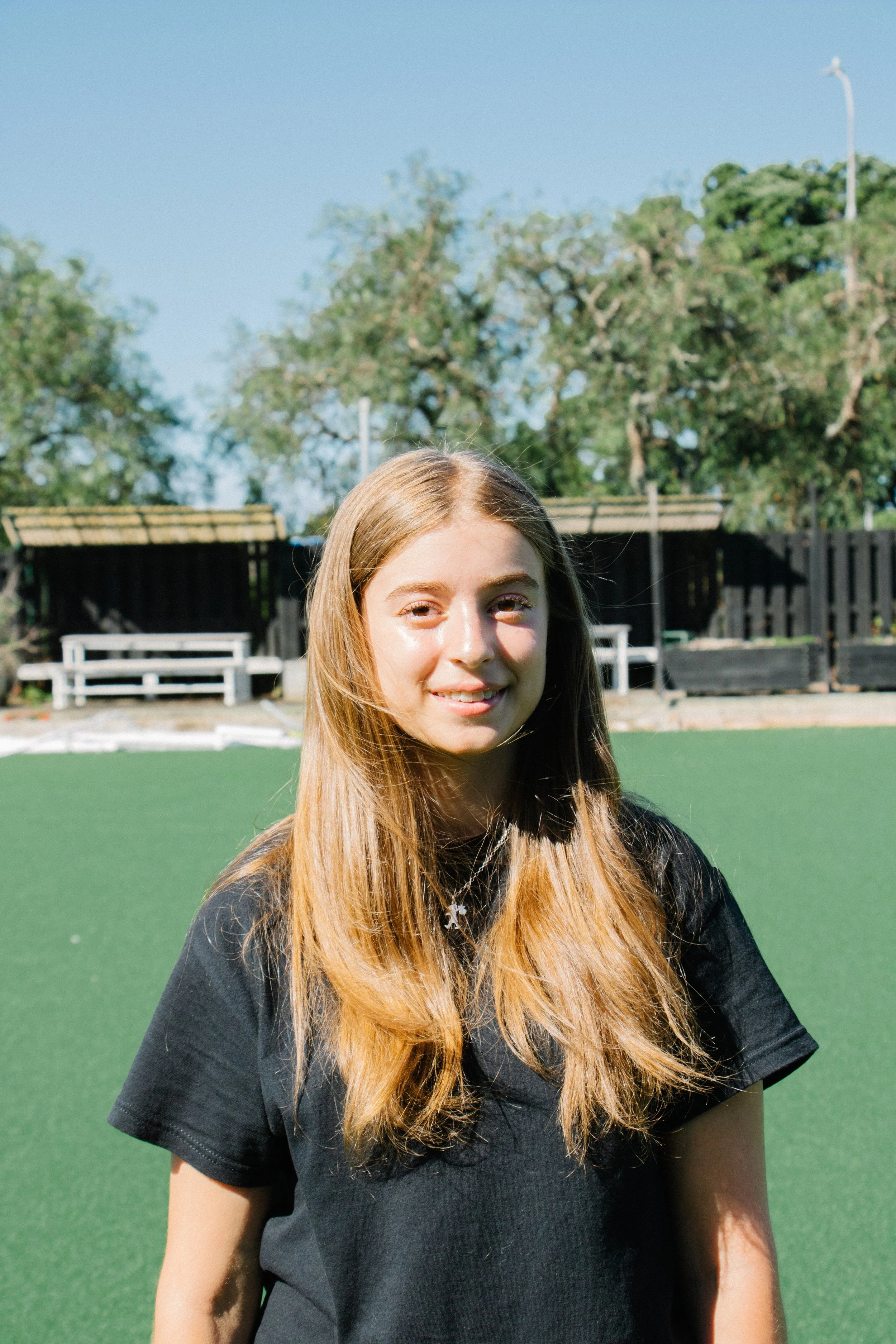 A young girl with long blonde hair smiling outdoors on a sunny day, standing on a green field with trees and benches in the background.