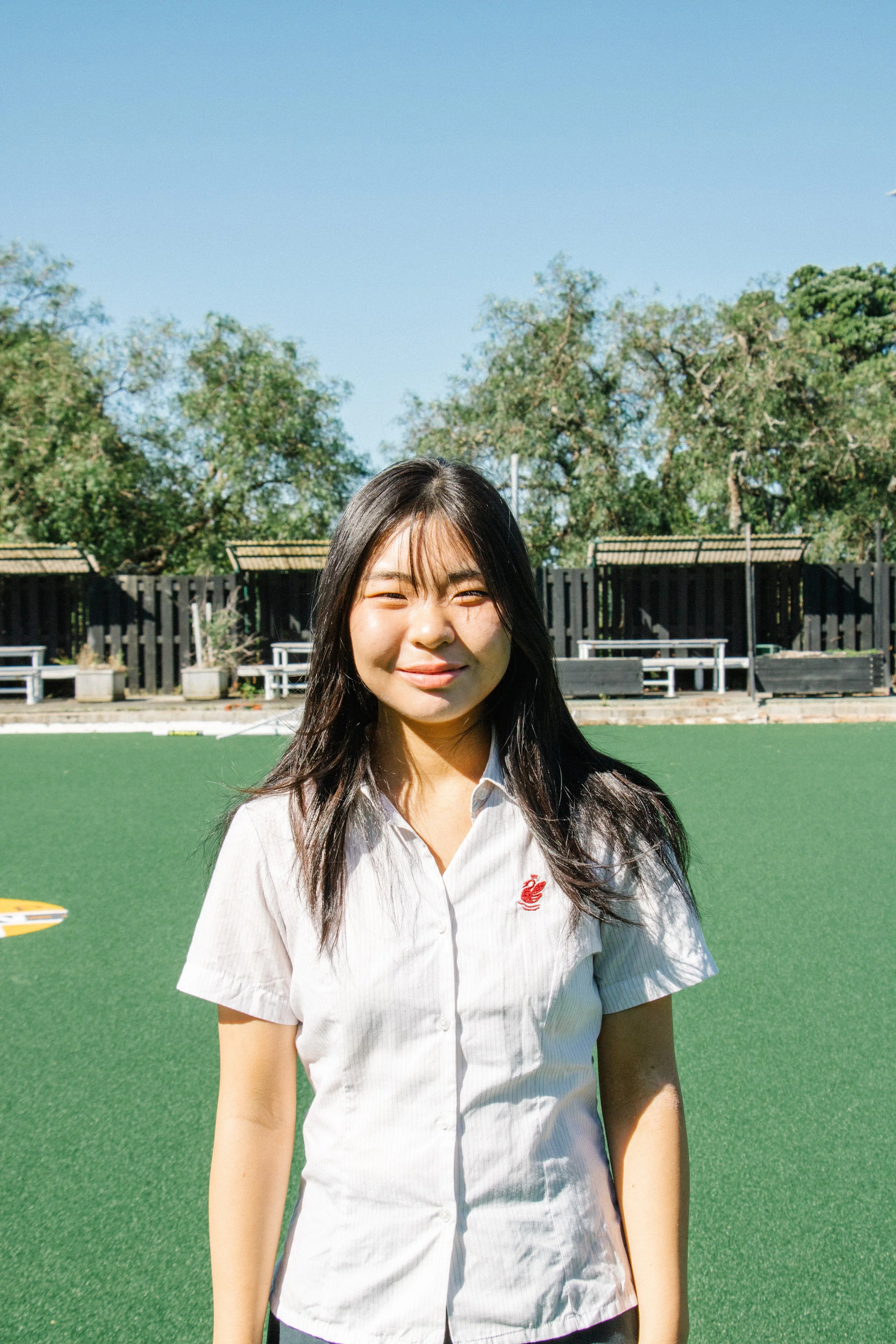 Young woman with long dark hair standing outdoors on a sunny day, wearing a white short-sleeved shirt with a small red embroidered logo, on a green sports field with trees and a black fence in the background.