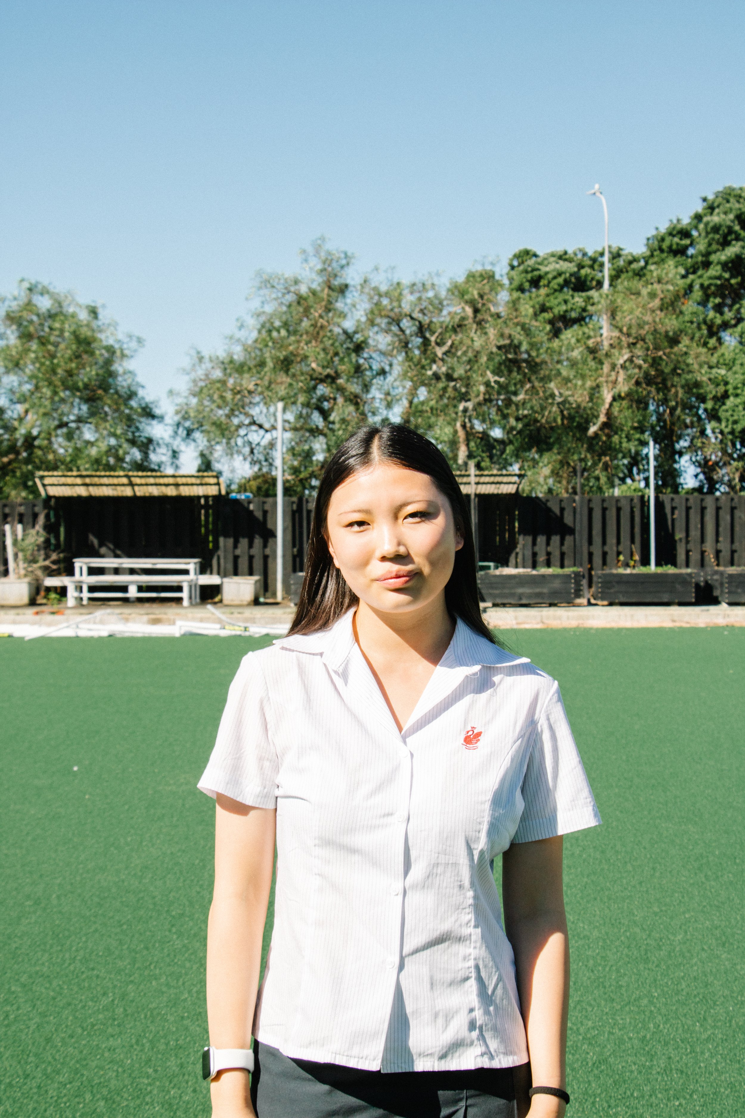 A young woman standing on a green outdoor field with trees, benches, and fence in the background, wearing a white short-sleeved shirt with a small red emblem and a smartwatch.