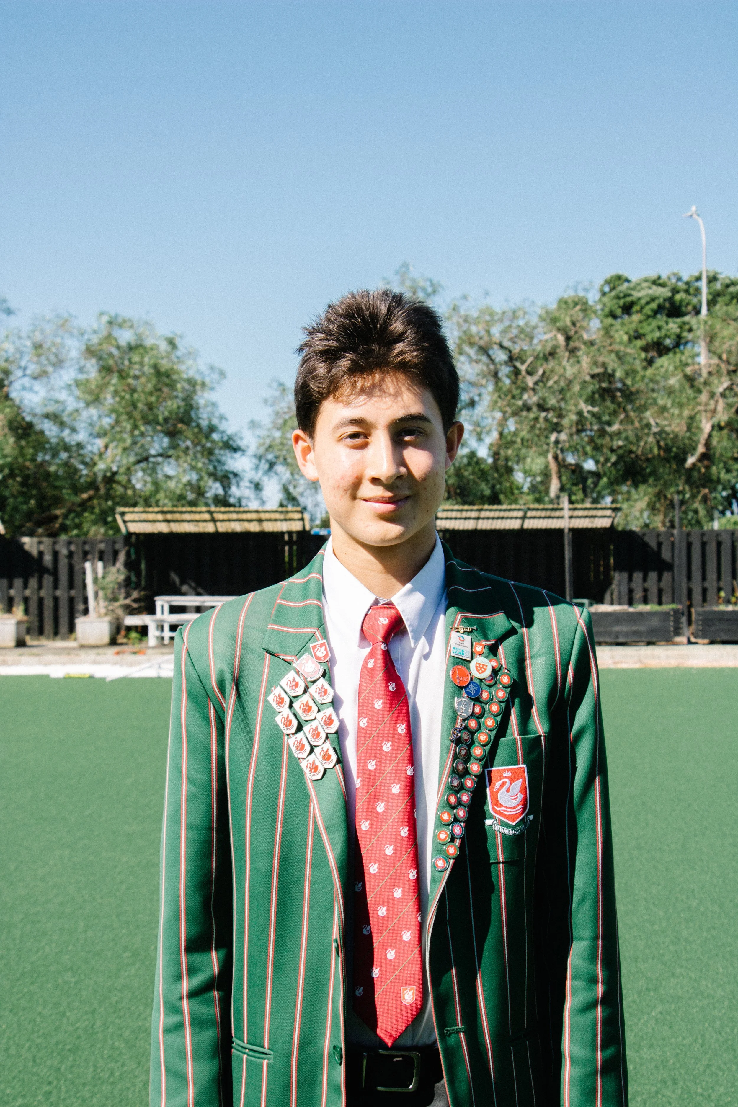 A young person standing outdoors on a grassy field, wearing a green blazer with white and red vertical stripes, a white shirt, a red tie, and an array of pins and badges on their blazer. The background includes trees, a black fence, and a clear blue sky.