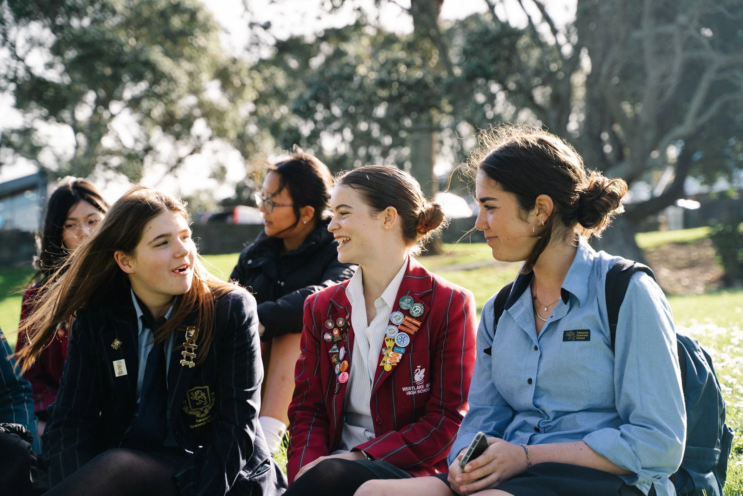 A group of teenage girls sitting outdoors on grass, smiling and chatting, with trees in the background.