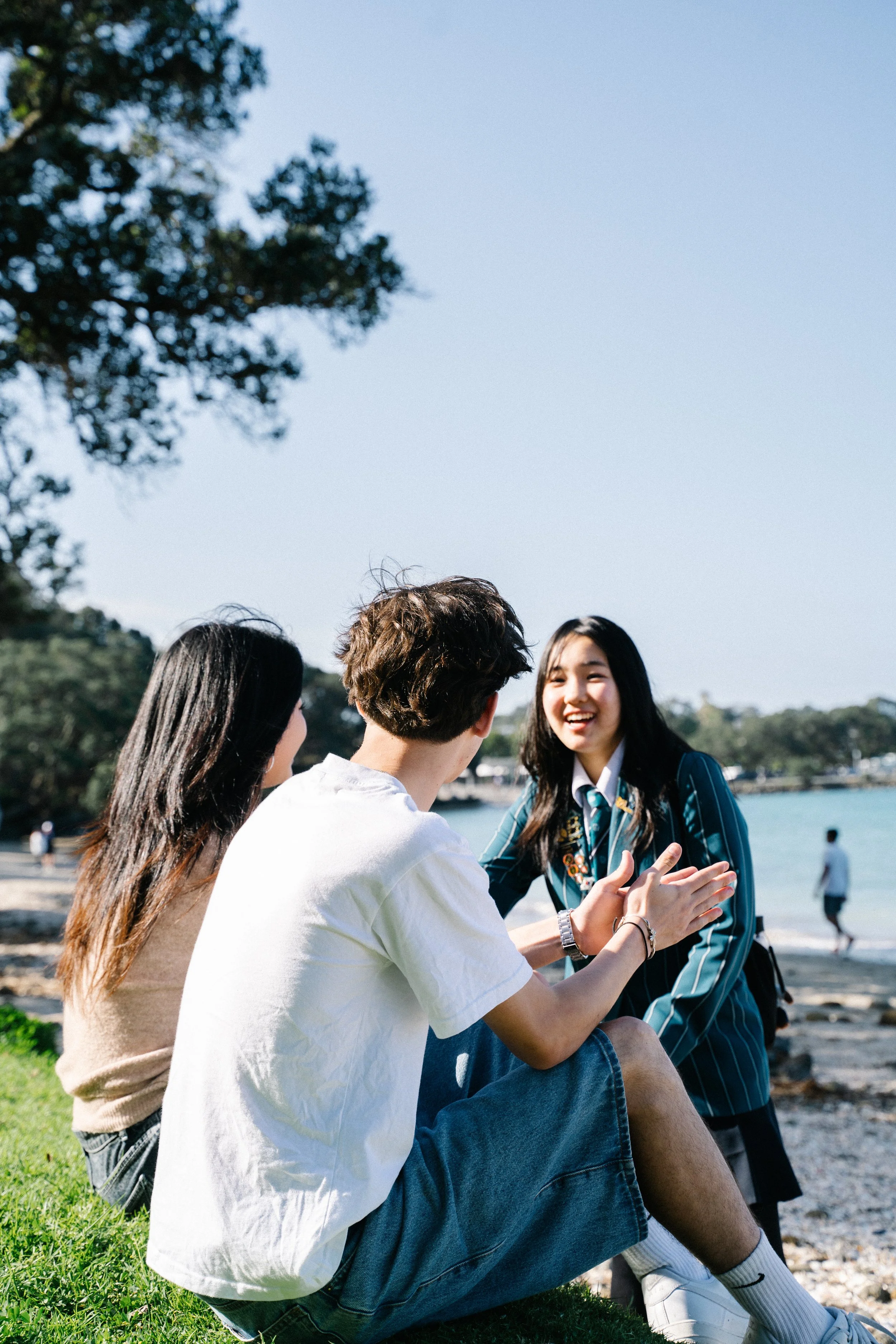 Three young people talking and laughing on a grassy area near a beach.