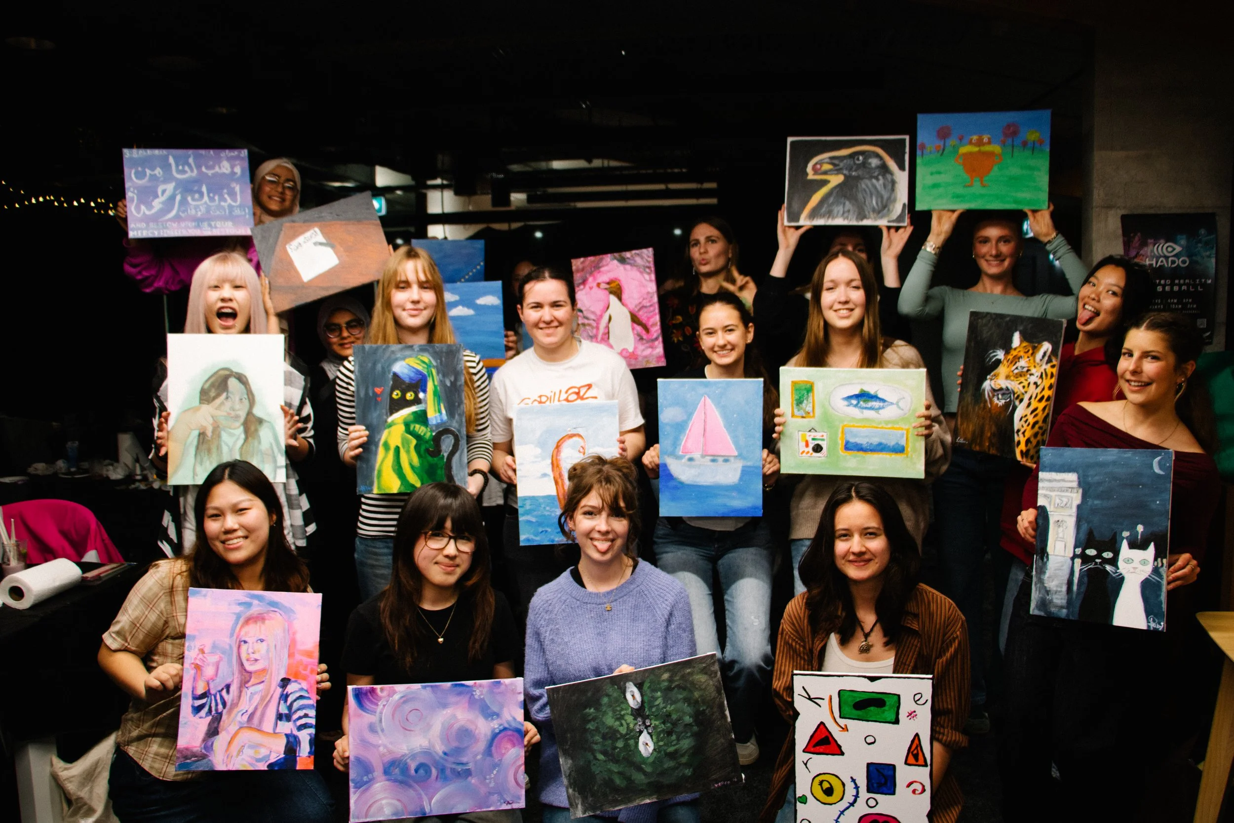Group of young women holding individual paintings at an art event or gallery.