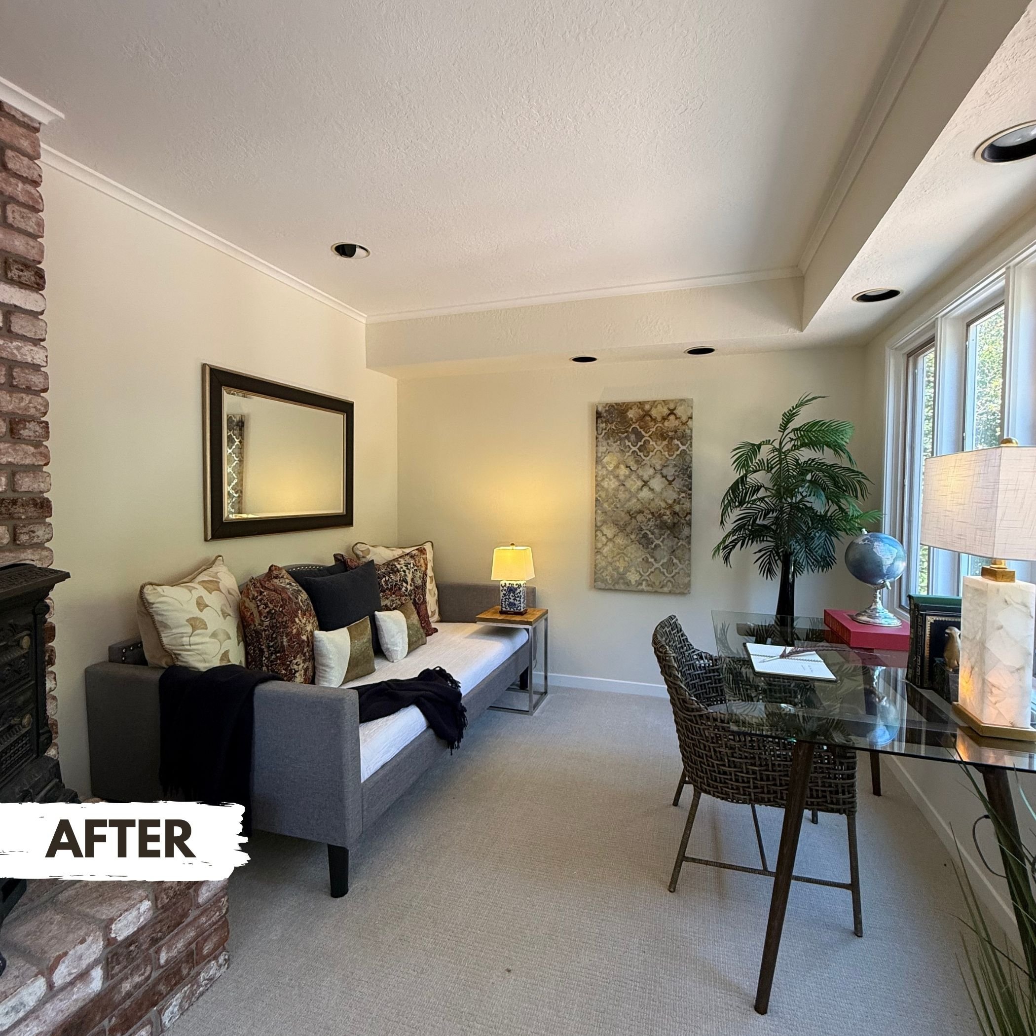 Updated living room with a gray sofa decorated with multiple patterned and solid throw pillows, a small side table with a lamp, a wicker chair with a glass-top table and a large green potted plant near a window, and a wall-mounted artwork.