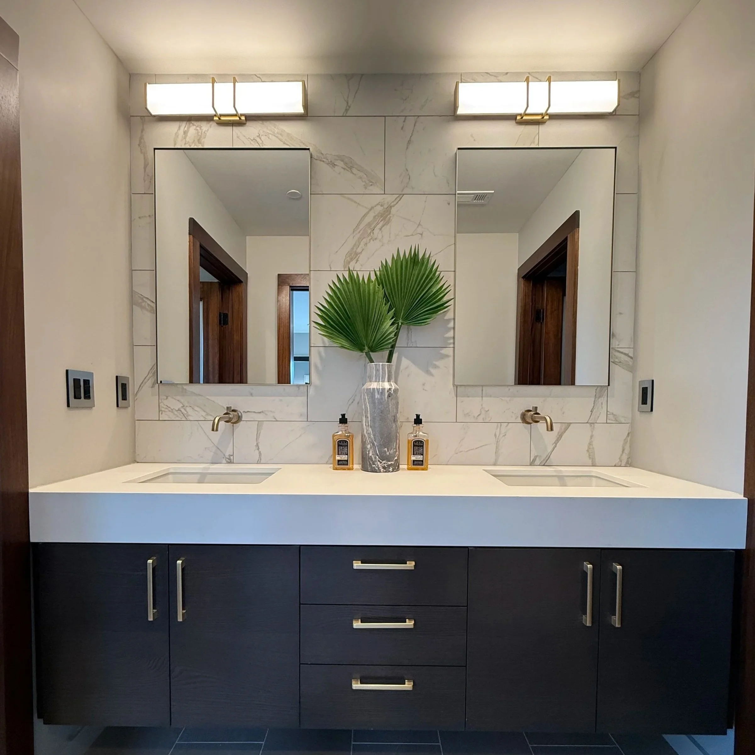 Double vanity bathroom with a marble backsplash, two rectangular mirrors, modern wall-mounted faucets, a large vase with green palm leaves, two soap dispensers, and dark cabinet with silver handles.