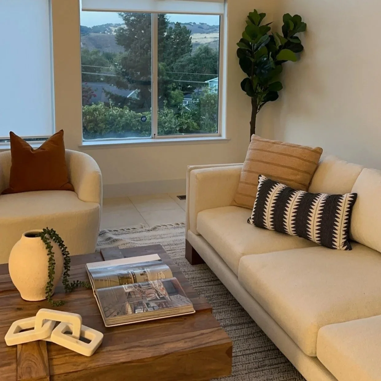 Living room with cream-colored sofa and armchair, decorated with brown, striped, and black-and-white pillows. A wooden coffee table holds an open magazine, a decorative white link chain, and a white vase with greenery. A large window shows an outdoor