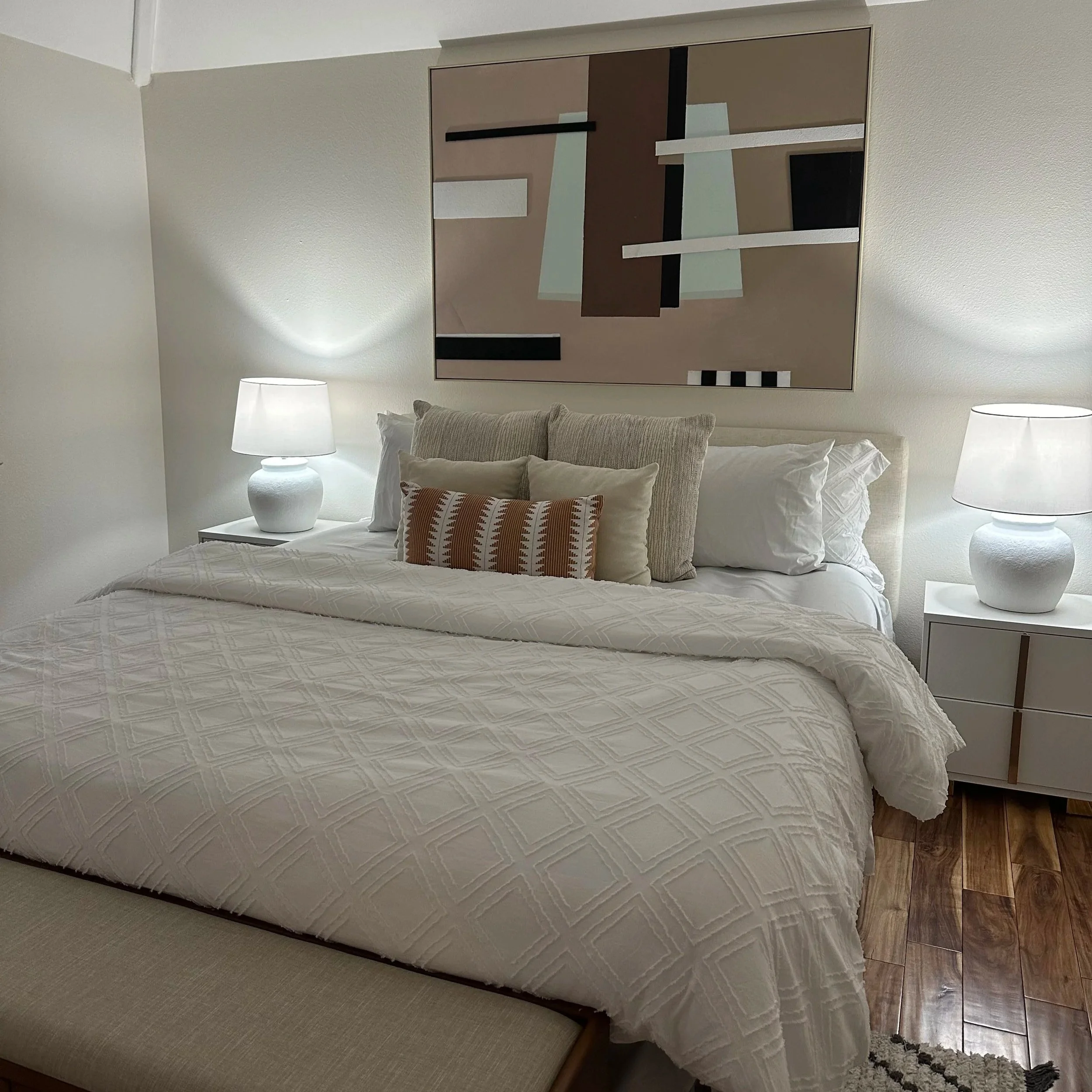 A neatly made bed with white bedding, four decorative pillows, and two bedside tables with matching white lamps in a bedroom with white walls and a wooden floor.