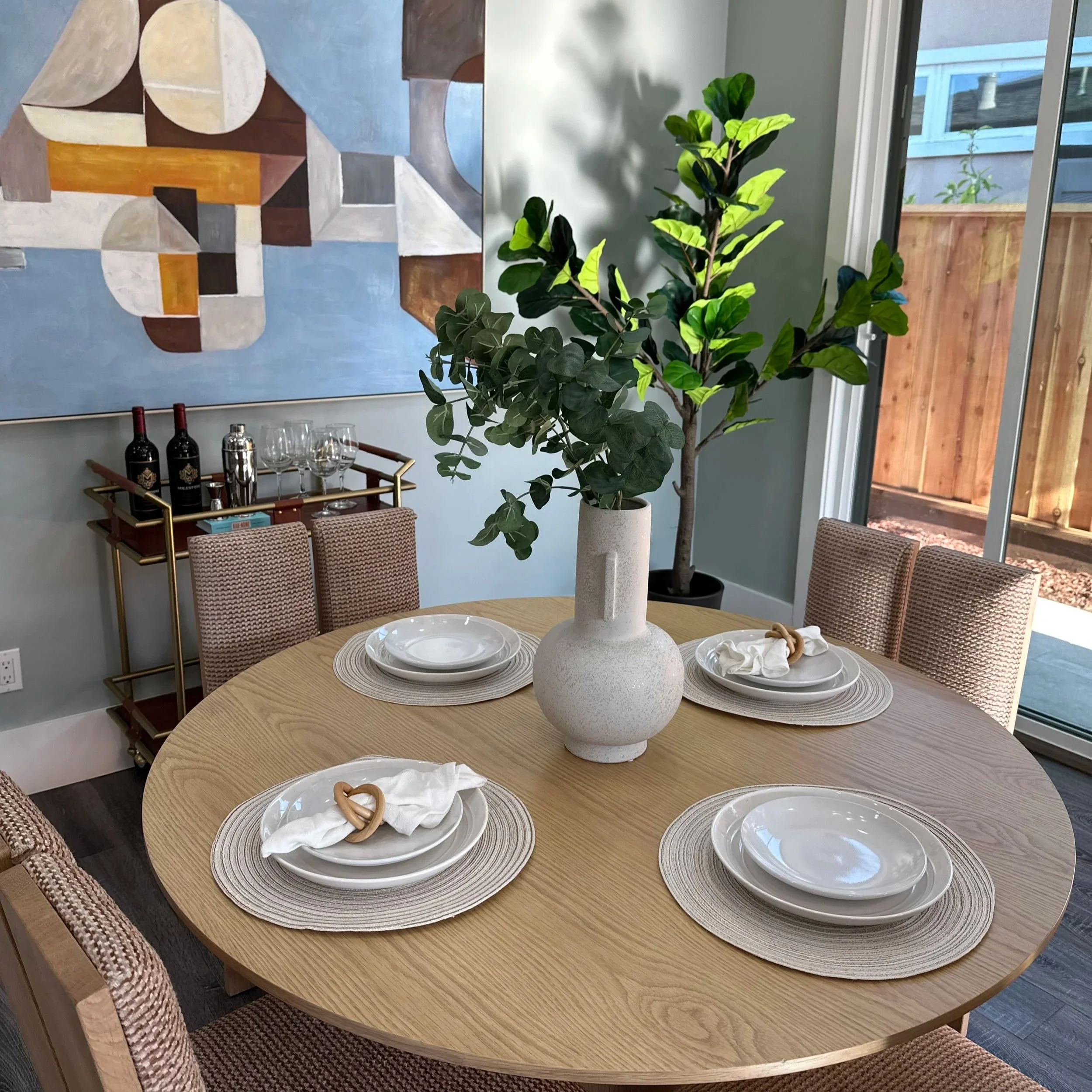 Round wooden dining table set with four place settings, each with a white plate, bowl, beige woven placemat, and cloth napkin with a wooden napkin ring. A large white textured vase with green leafy branches is in the center of the table. In the backg
