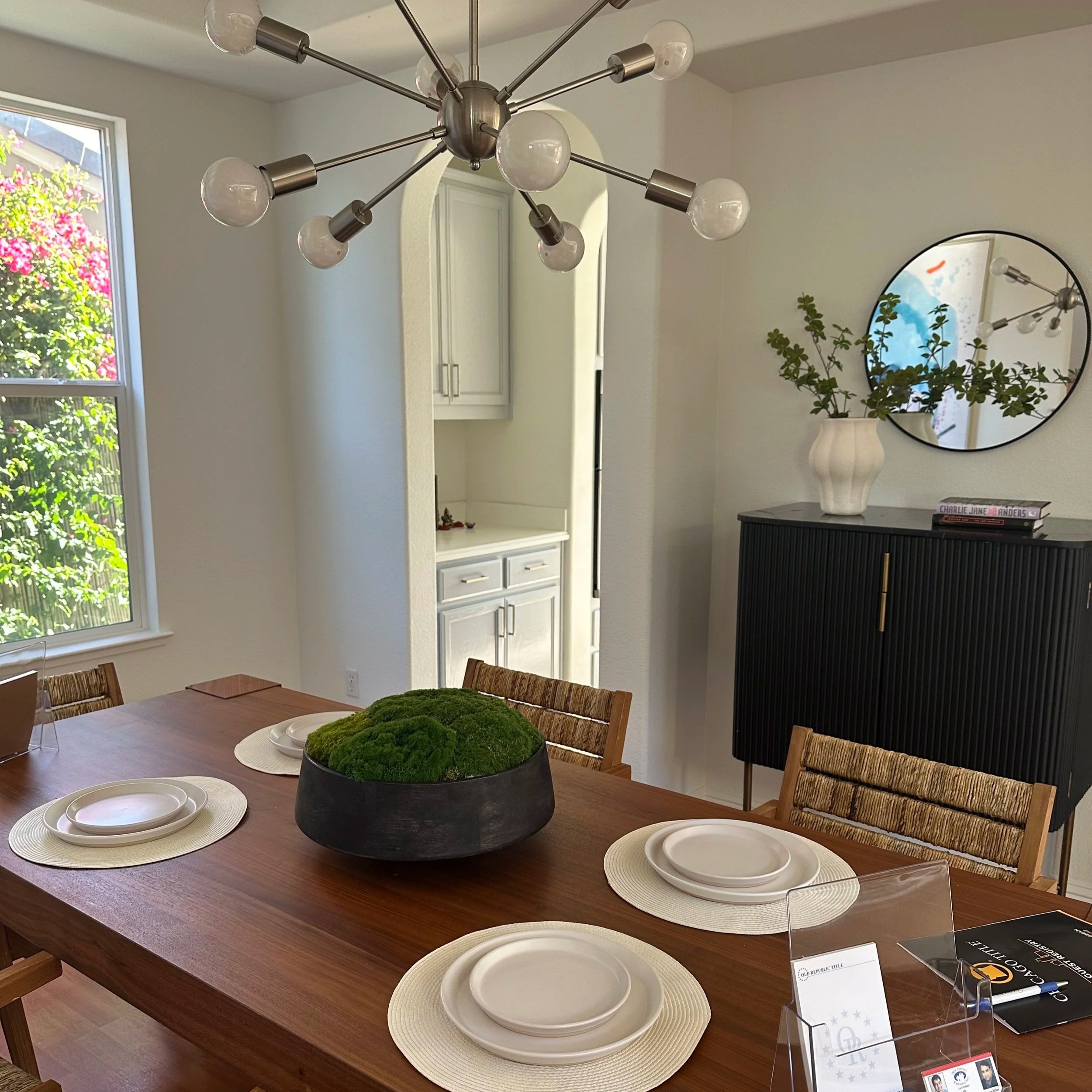 A dining room with a wooden table set with four white plates on woven placemats. A large round black bowl with green moss is in the center of the table. There are wooden chairs with striped upholstery around the table. A modern chandelier with multip