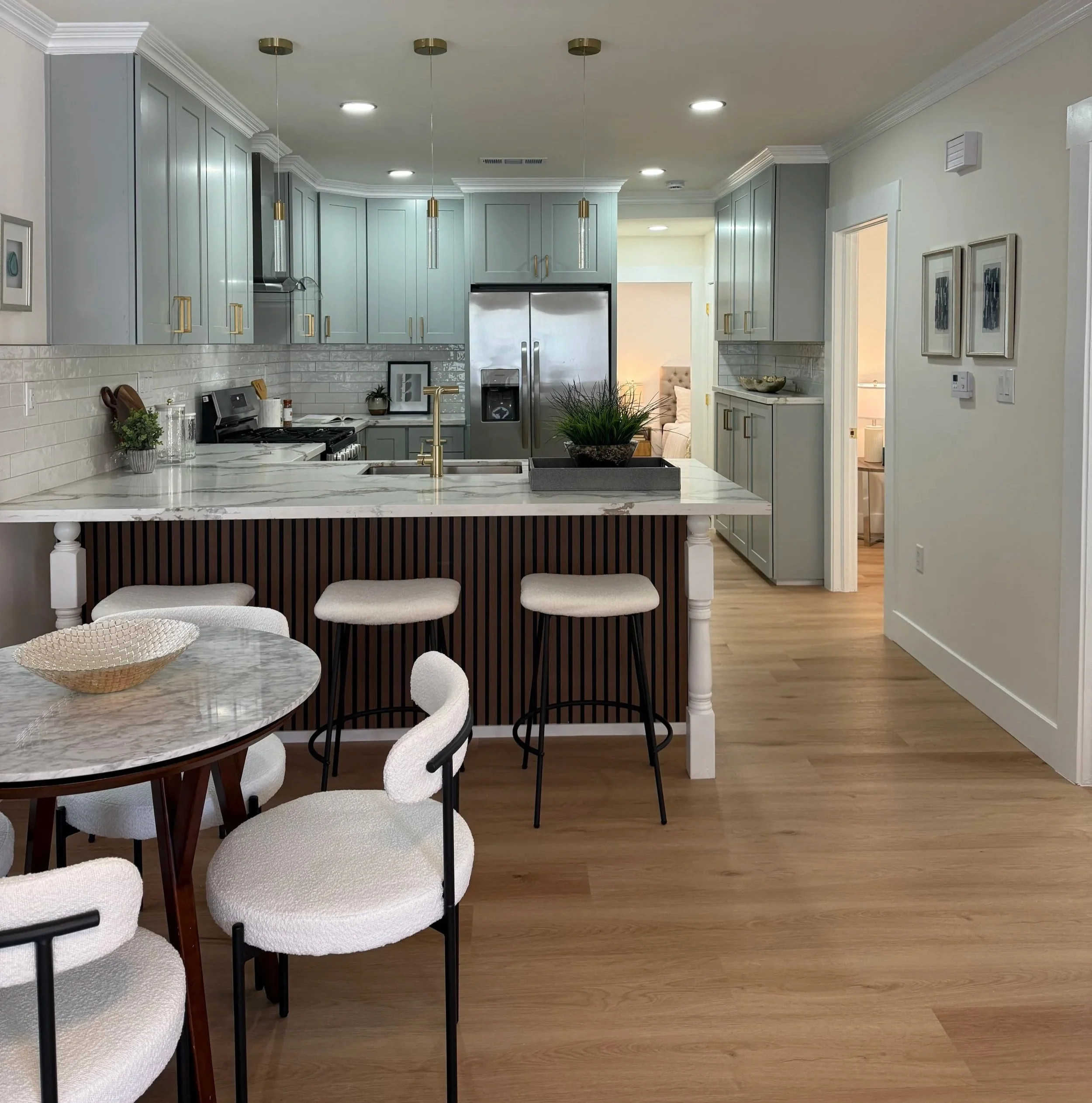 Modern kitchen with light blue cabinets, white marble countertops, white brick backsplash, stainless steel refrigerator, and a kitchen island with a planter on top. Light-colored hardwood floor and surrounding dining area.