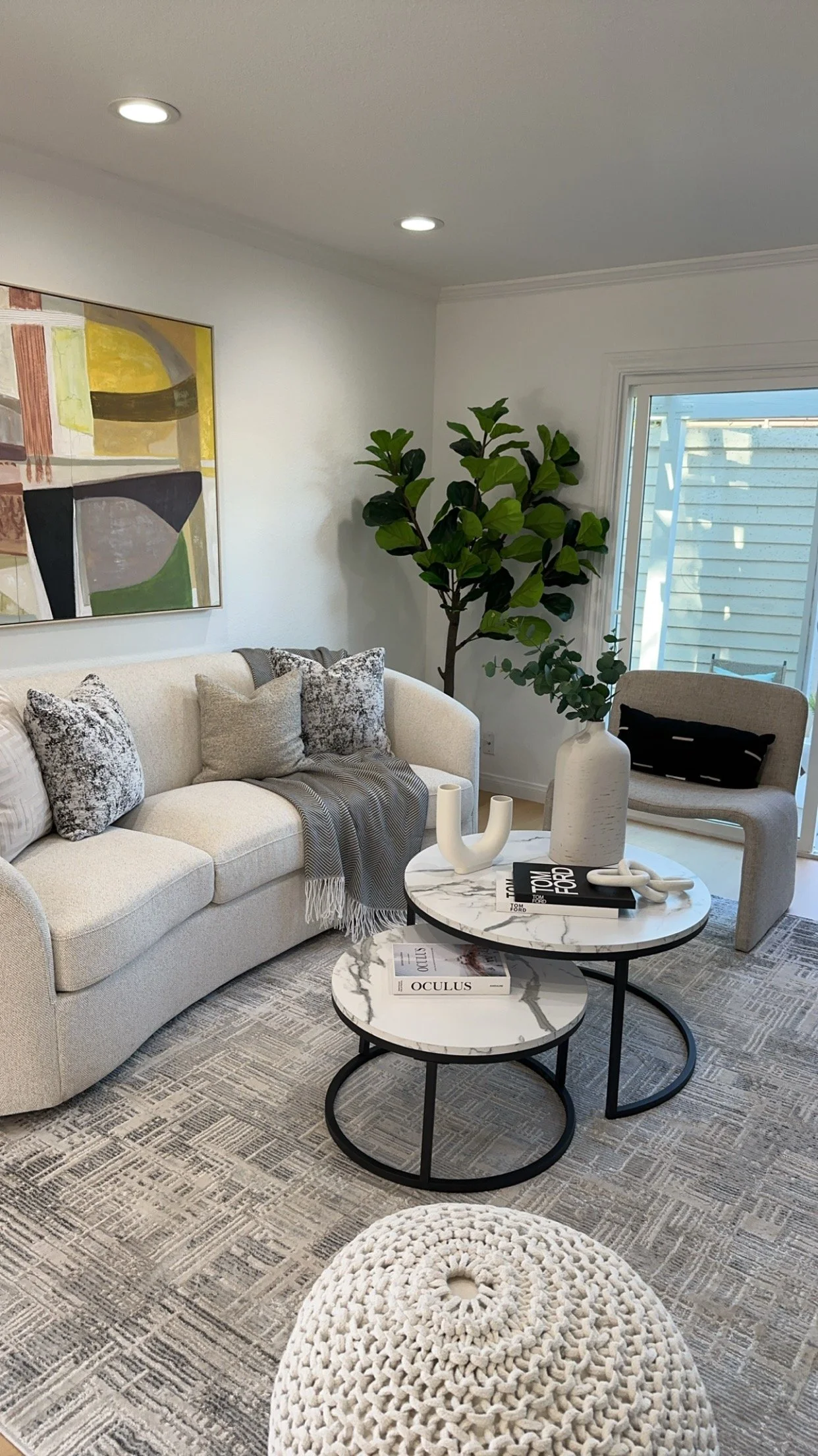 Modern living room with a white sofa, abstract art on the wall, green potted plant, sliding glass door, beige armchair with a black pillow, layered round marble-top coffee tables with decorative vases and books, and a textured floor mat.