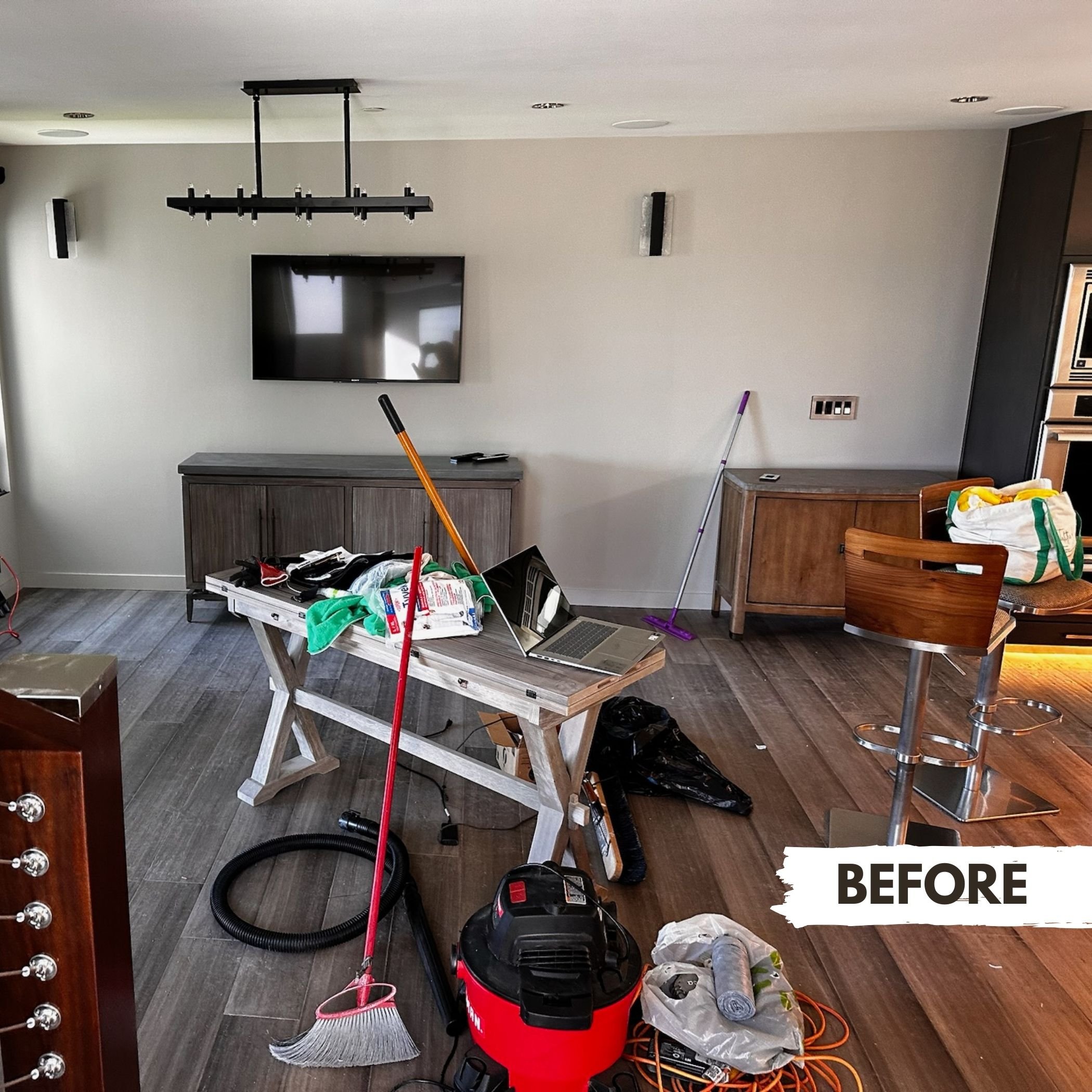 Living room under cleaning, with cleaning tools, vacuum cleaner, broom, and dustpan on the floor, a laptop on a wooden table, a TV mounted on the wall, and furniture including a bar stool, sideboard, and cabinets.