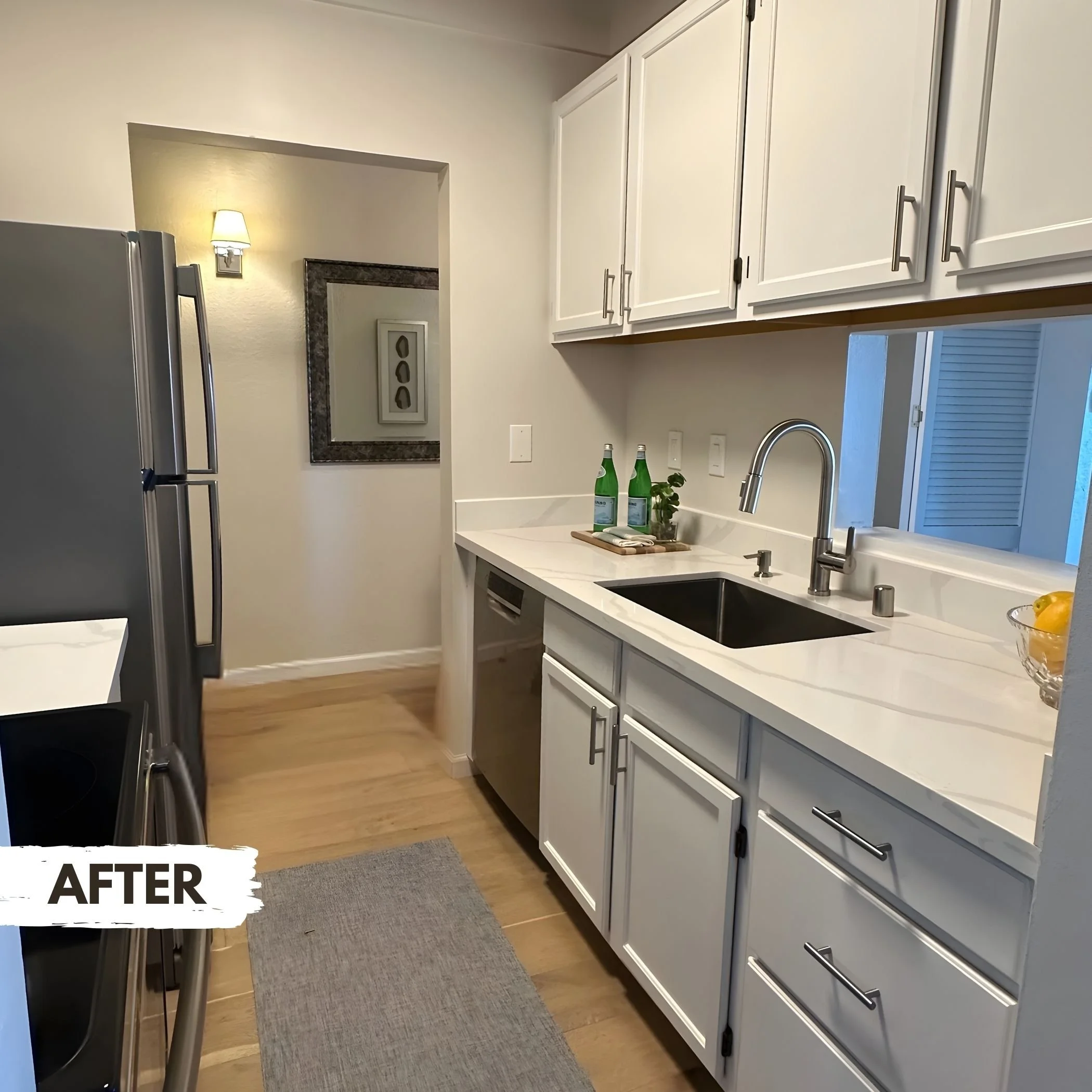 Modern kitchen with white cabinets, a black sink, stainless steel dishwasher, and wooden flooring. Two bottles of sparkling water, a small plant, and a bowl of lemons are on the countertop.