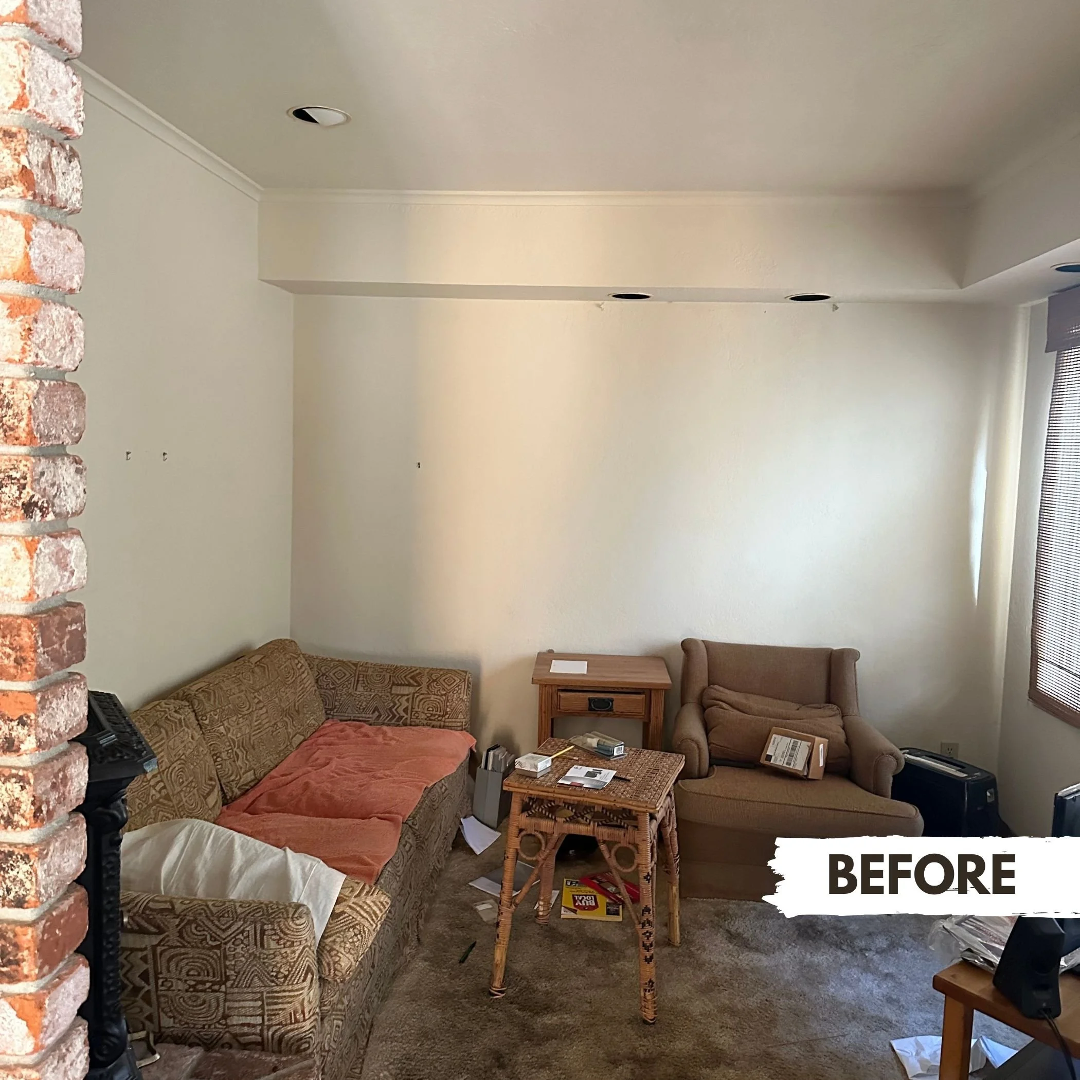Living room with beige walls, a patterned sofa with a pinkish throw, a single armchair, a small wooden side table, a small wooden table in front of the sofa, some scattered papers and objects, and a window with partially closed blinds.