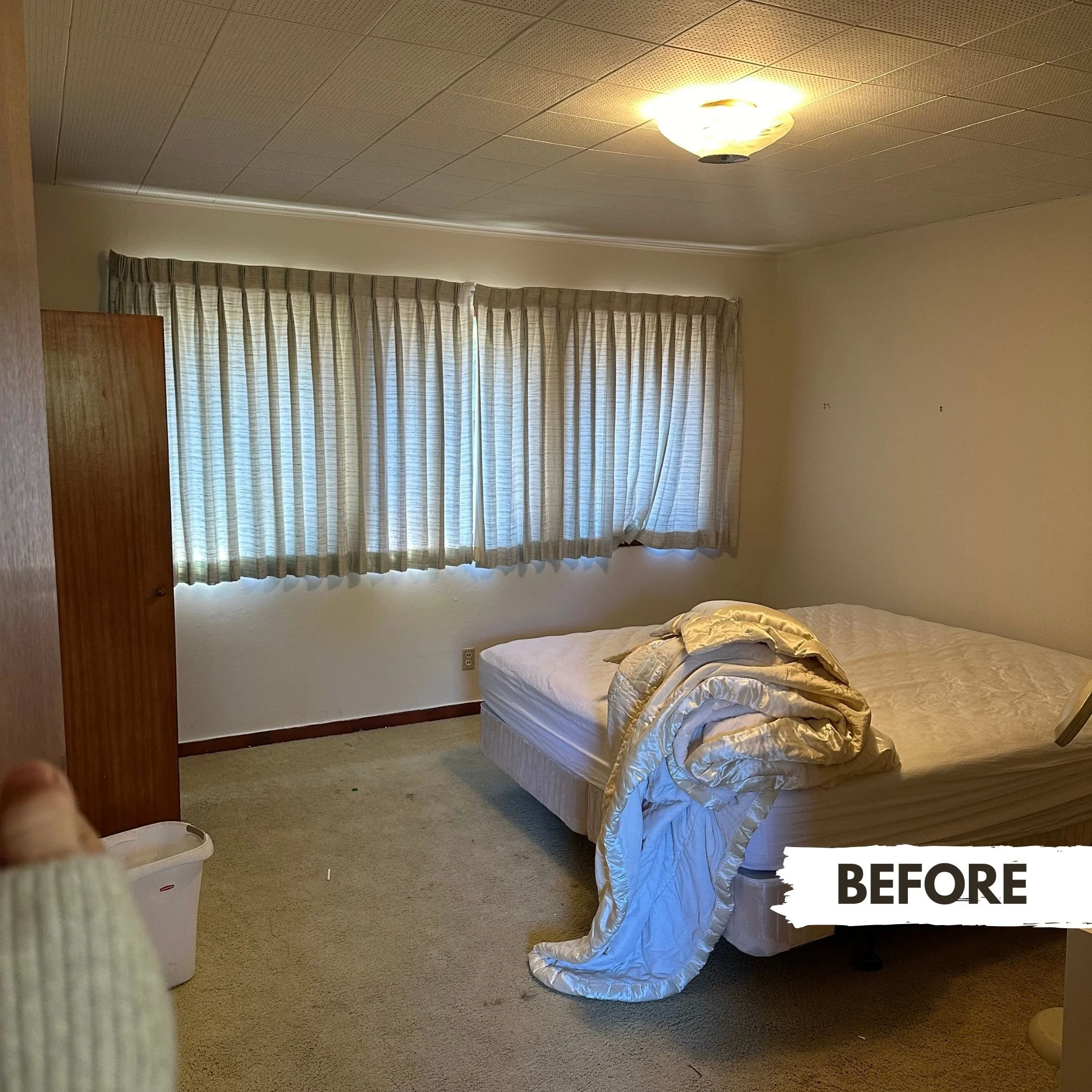 An uncluttered bedroom with a bed without linens, a large window with closed curtains, and a ceiling light fixture.