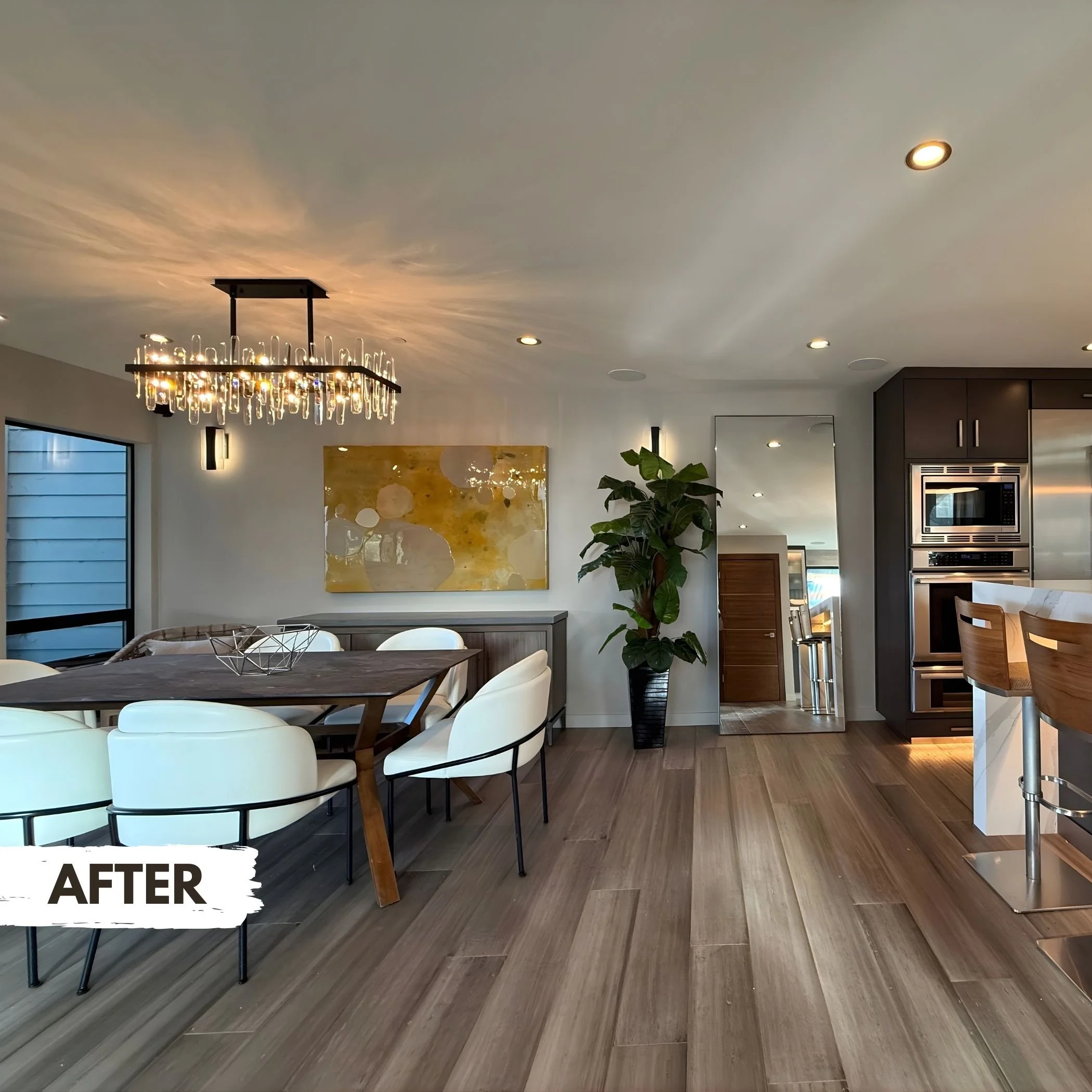 Modern dining area with a wooden table surrounded by white chairs, a large abstract painting on the wall, a tall mirror, and a big potted plant. The ceiling has a decorative chandelier, and the space is bright with recessed lighting. A kitchen with m