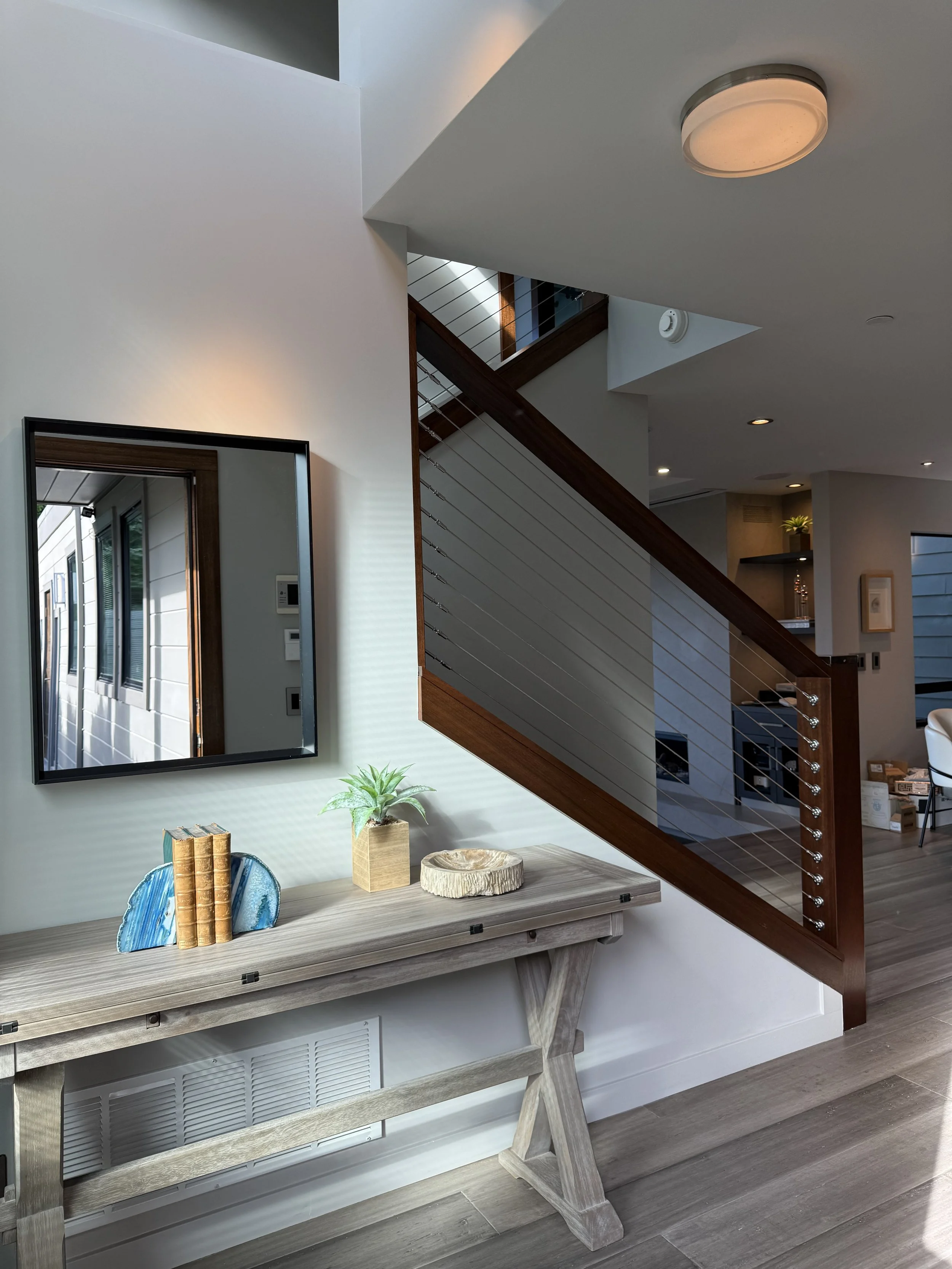 Interior of a modern home with a wooden console table, decorative books, a green plant in a wooden pot, a wooden bowl, a square mirror, a staircase with glass and wood railing, and ceiling lights.