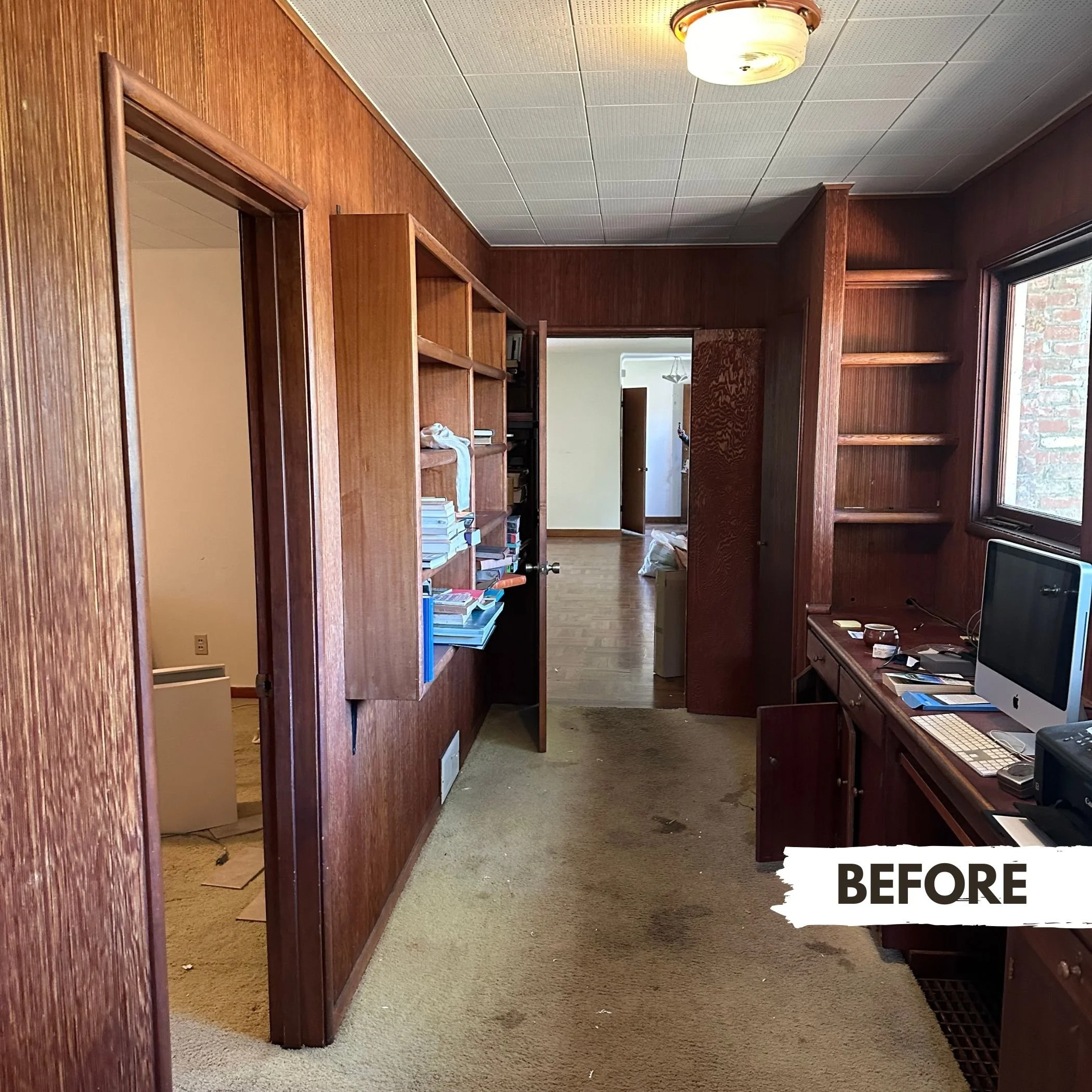 A cluttered home office with brown wood-paneled walls, a window, and a desk with a computer, keyboard, and various items. The carpet is stained, and there are open shelves with books and papers. An adjoining room with wood flooring is visible through