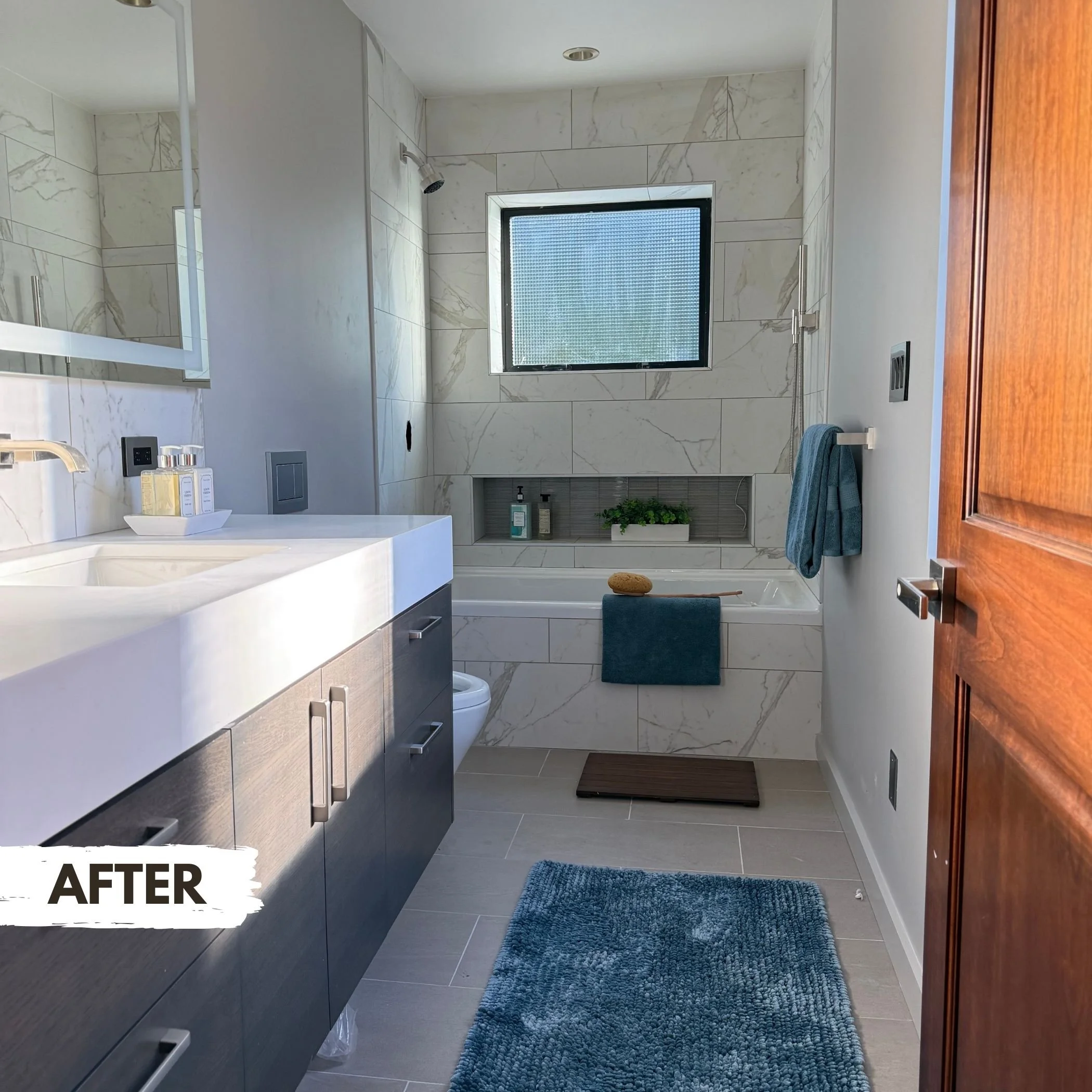 Modern bathroom with marble walls, a white bathtub with blue towels and green plants, a large mirror, gray vanity, and wooden door.