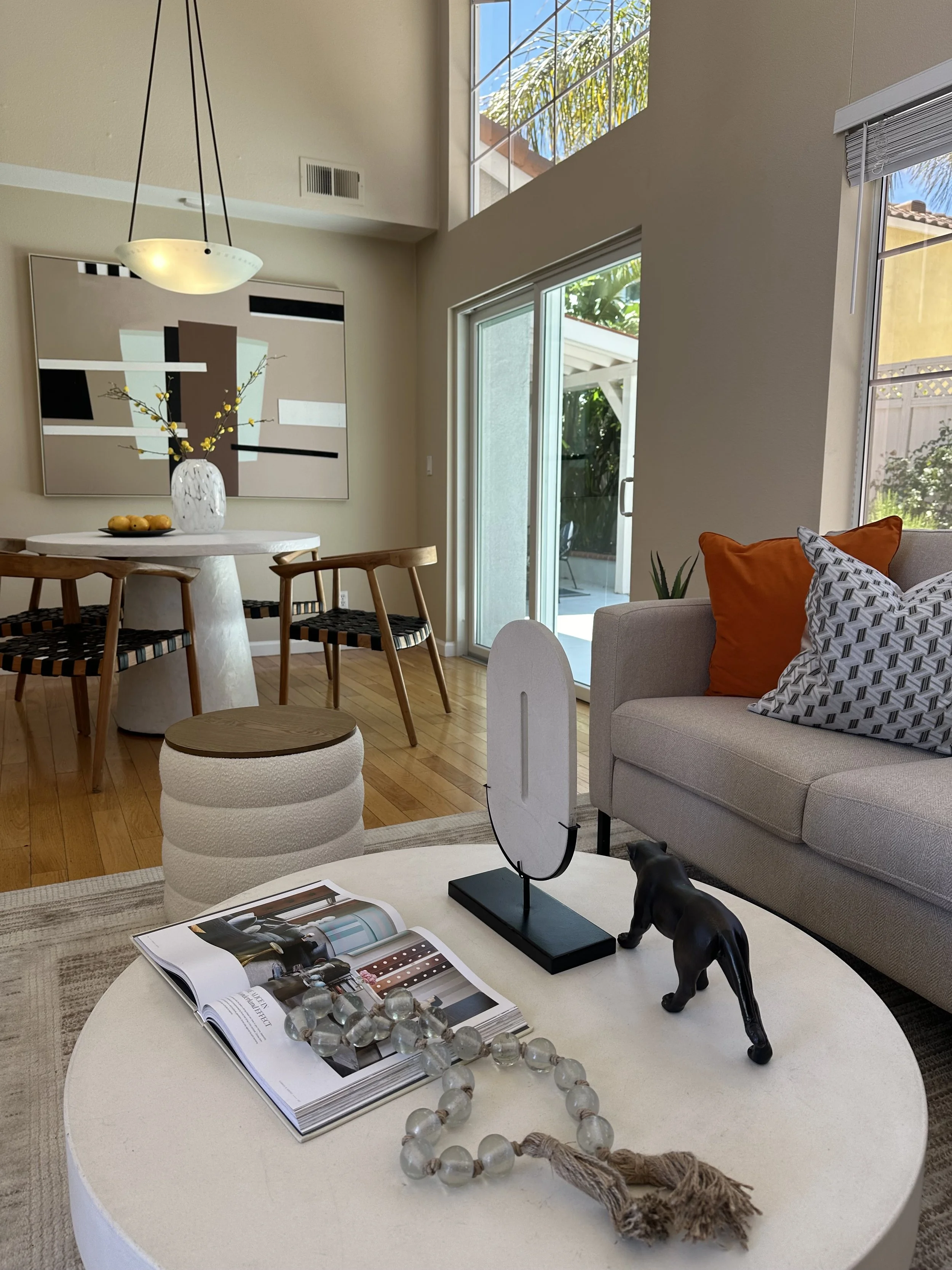 Living room with a round white coffee table, a beige sofa with orange and patterned pillows, open magazine, decorative bookstand, glass bead necklace, black dog figurine, and a large abstract painting on the wall. In the background, a dining area wit