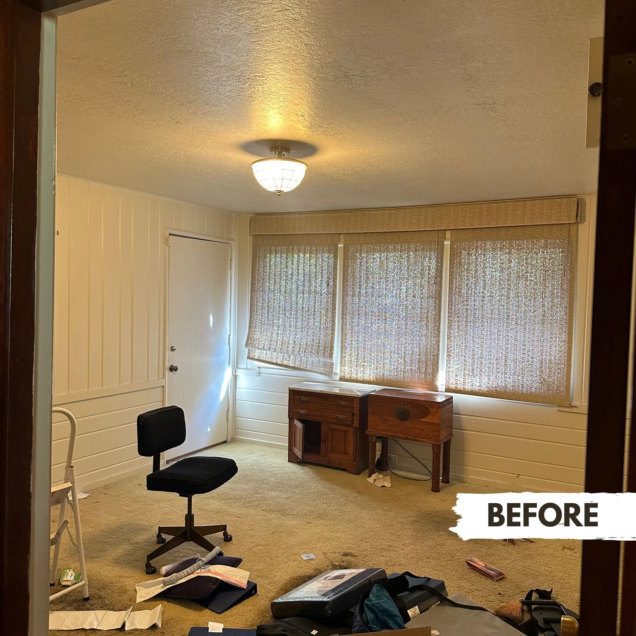 A cluttered room with beige carpet, a white door, a fabric window shade, and liners scattered on the floor. There is a black office chair and a small white step stool. Two wooden cabinets are positioned by the window, and a ceiling light fixture illu