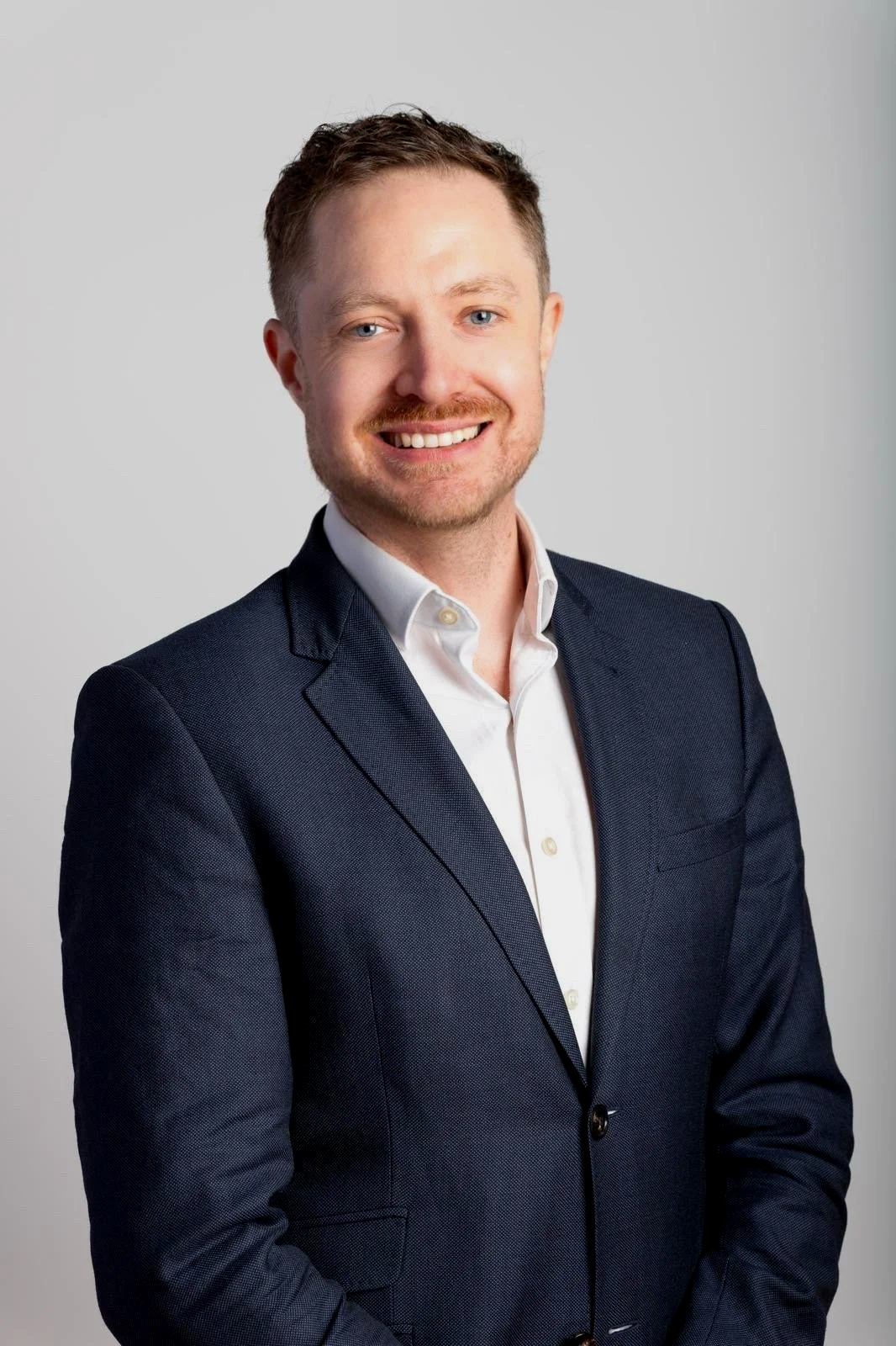 A professional man with short brown hair, blue eyes, and a mustache, wearing a dark suit jacket over a white shirt, smiling at the camera.